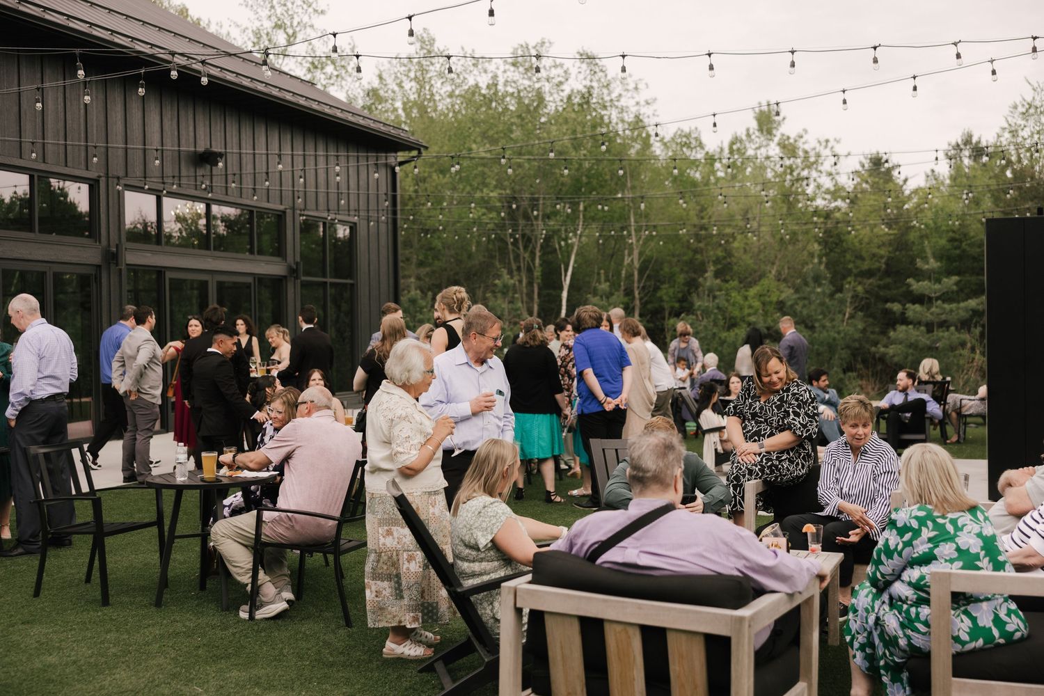 Guests mingle at an outdoor evening reception with string lights, dark barn venue, and lush greenery in the background.