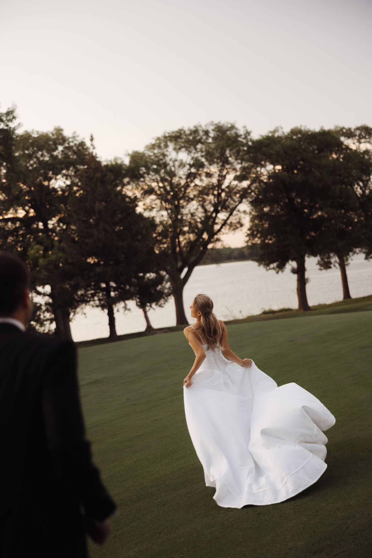 Bride in flowing white gown walks toward a serene lakeside lawn at golden hour, with groom watching in foreground.