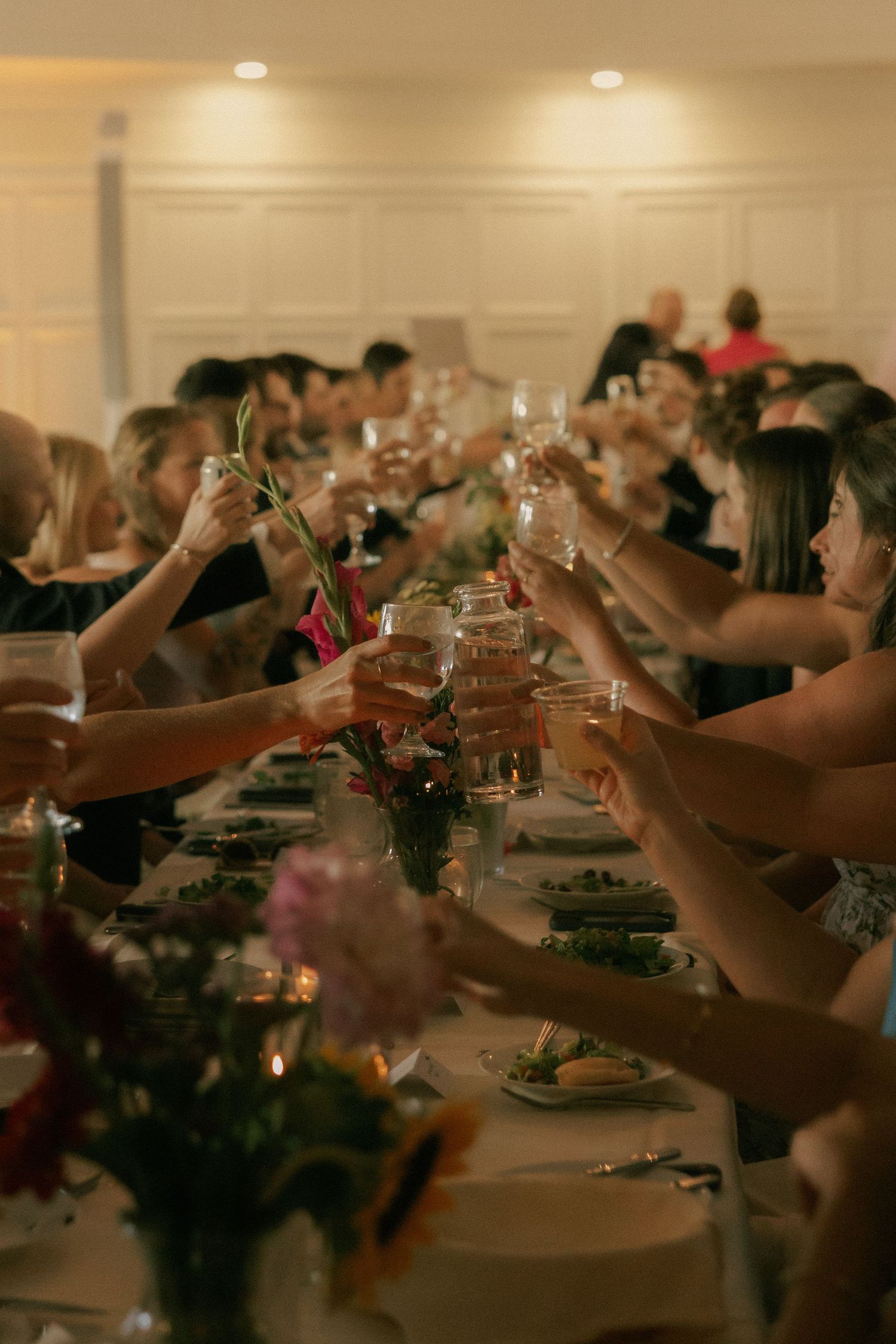 Guests raising glasses in a toast at a long dinner table decorated with flowers and candles at an elegant event.