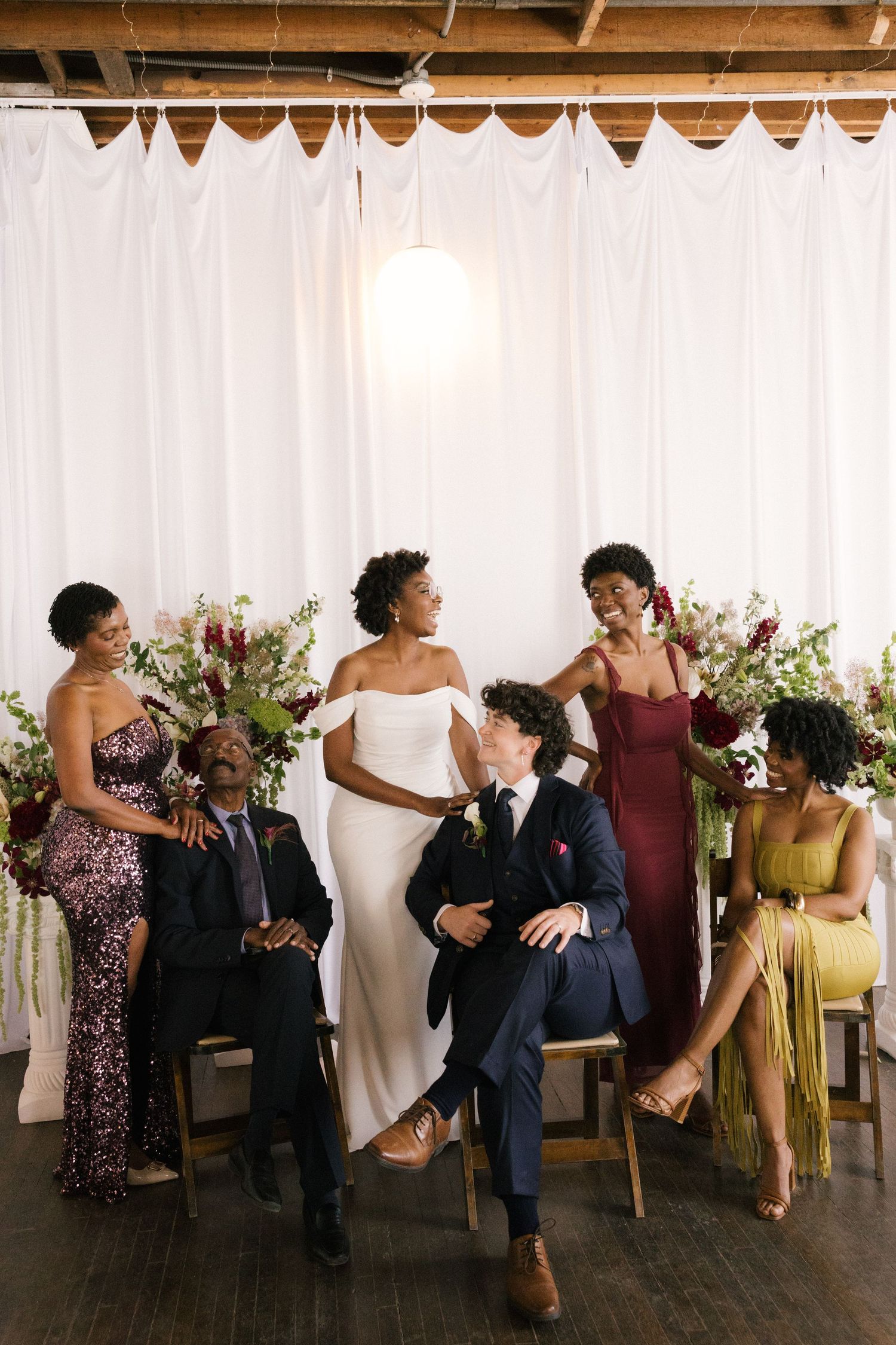 Bride in white gown poses with groom and wedding party in front of white drape backdrop with floral arrangements.