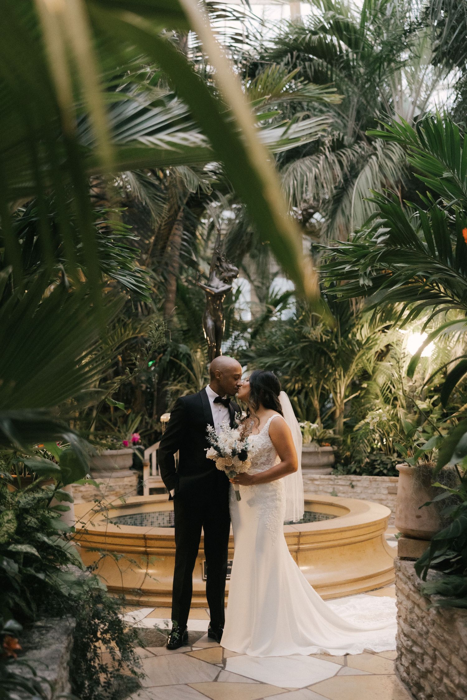 A couple shares an intimate moment during their wedding ceremony surrounded by lush tropical palm trees and greenery.