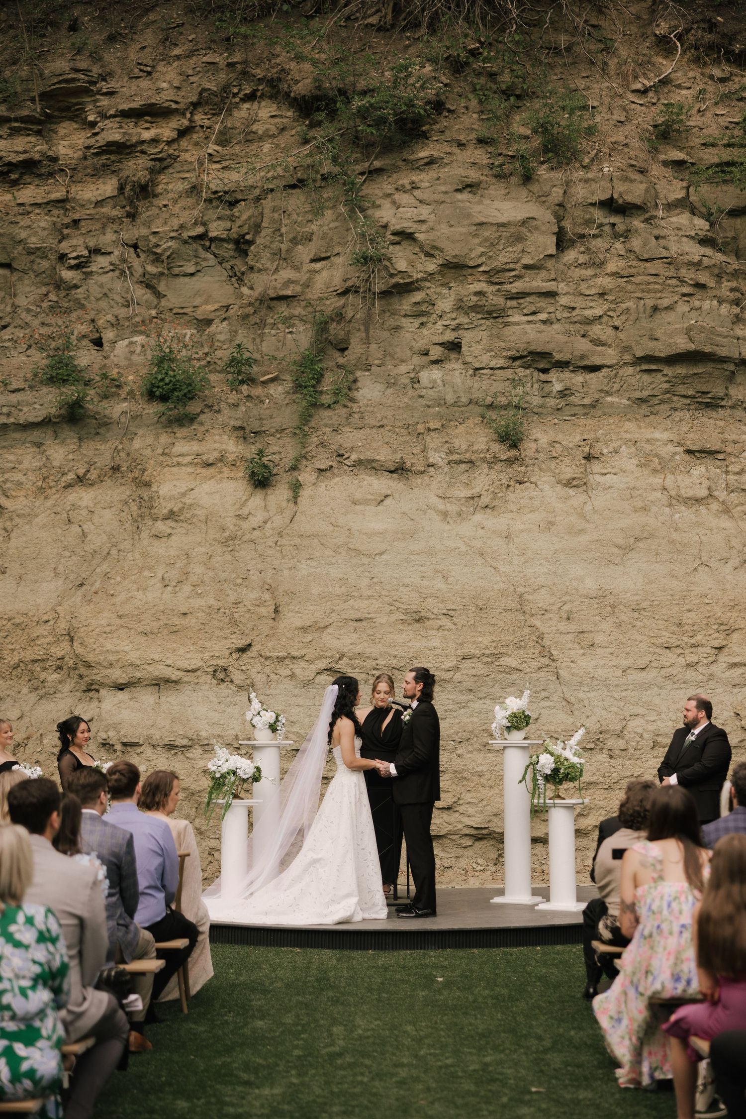 Bride and groom exchange vows on outdoor stage before stunning sandstone cliff backdrop with seated wedding guests.