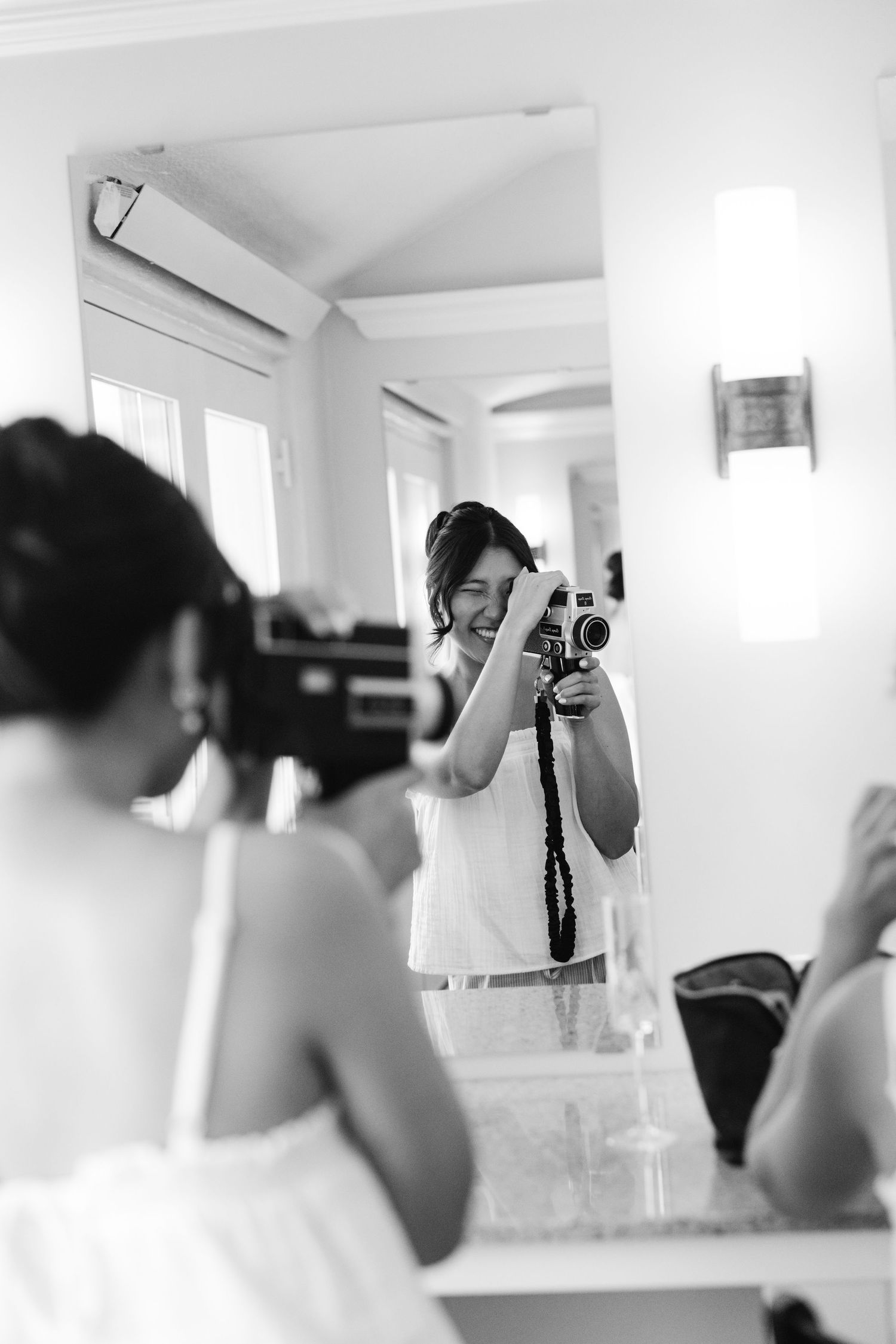 Black and white photo of a smiling woman photographing her reflection in a mirror with a vintage camera.