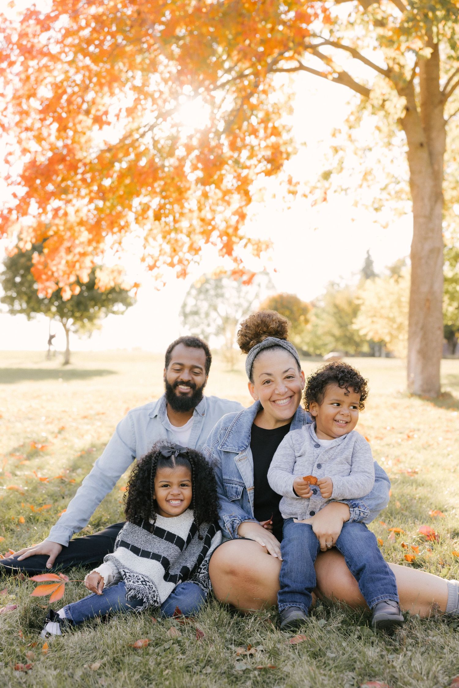 A family sits together on autumn grass beneath colorful fall foliage with sunlight streaming through the trees.