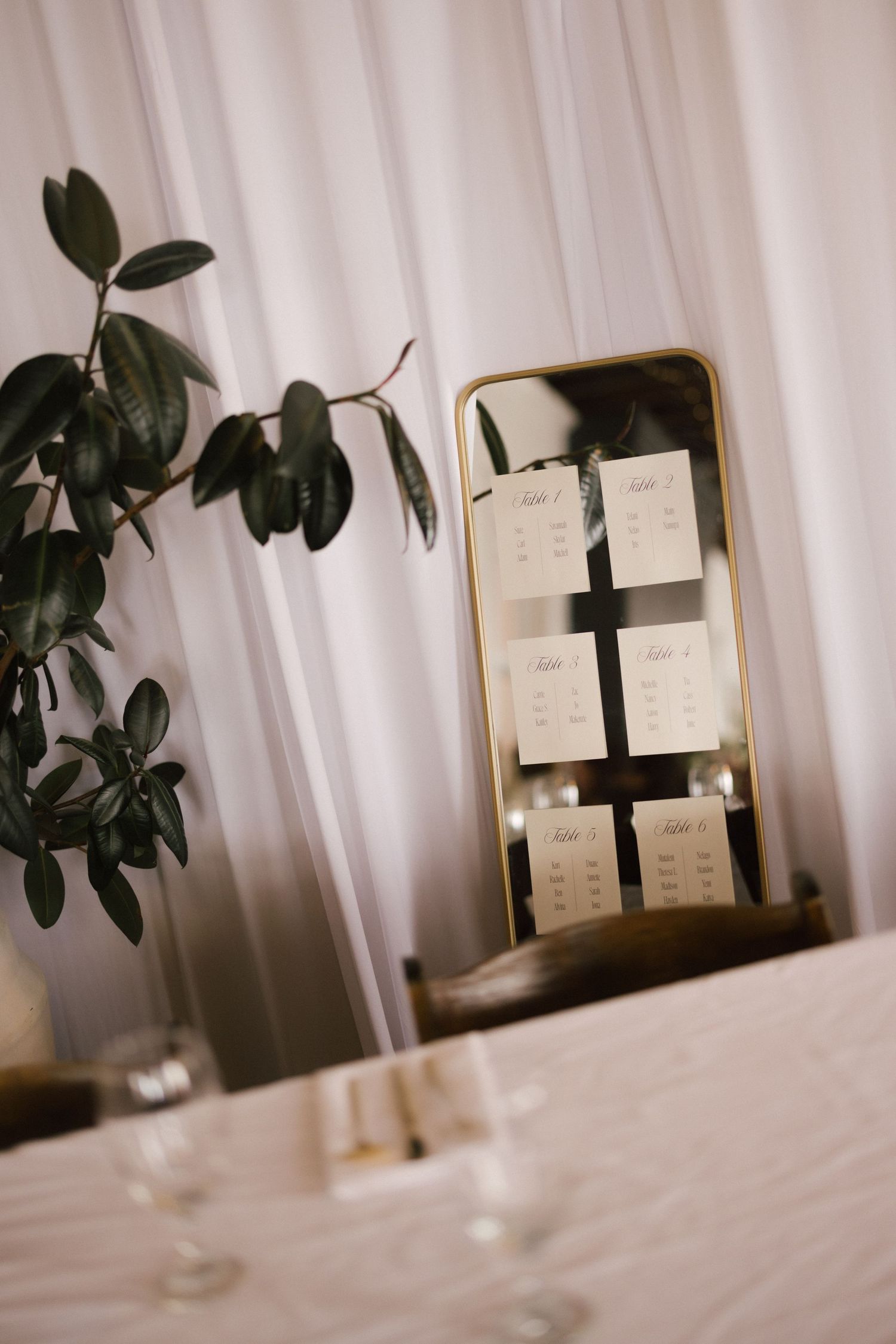 Elegant wedding seating chart displayed on a gold mirror frame with white draping and greenery.