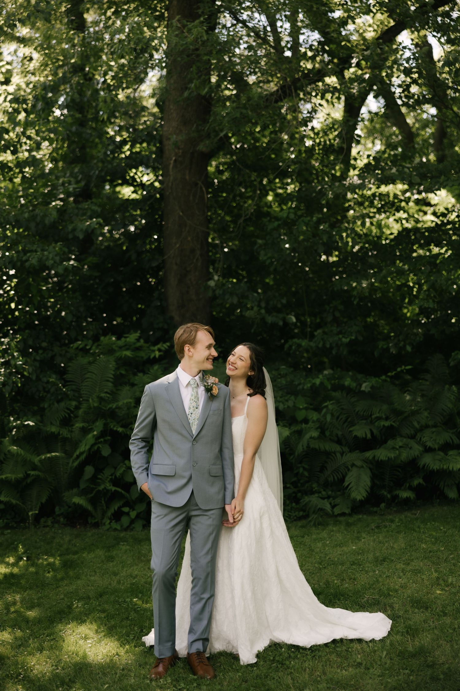 A bride and groom hold hands and smile in a lush green garden, she in a white gown, he in a gray suit.