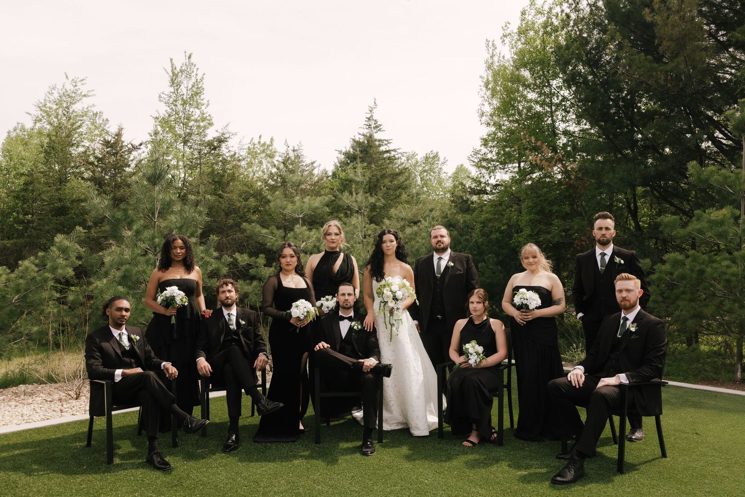 Wedding party in black attire poses outdoors on green lawn with trees, bride holding white bouquet in center.