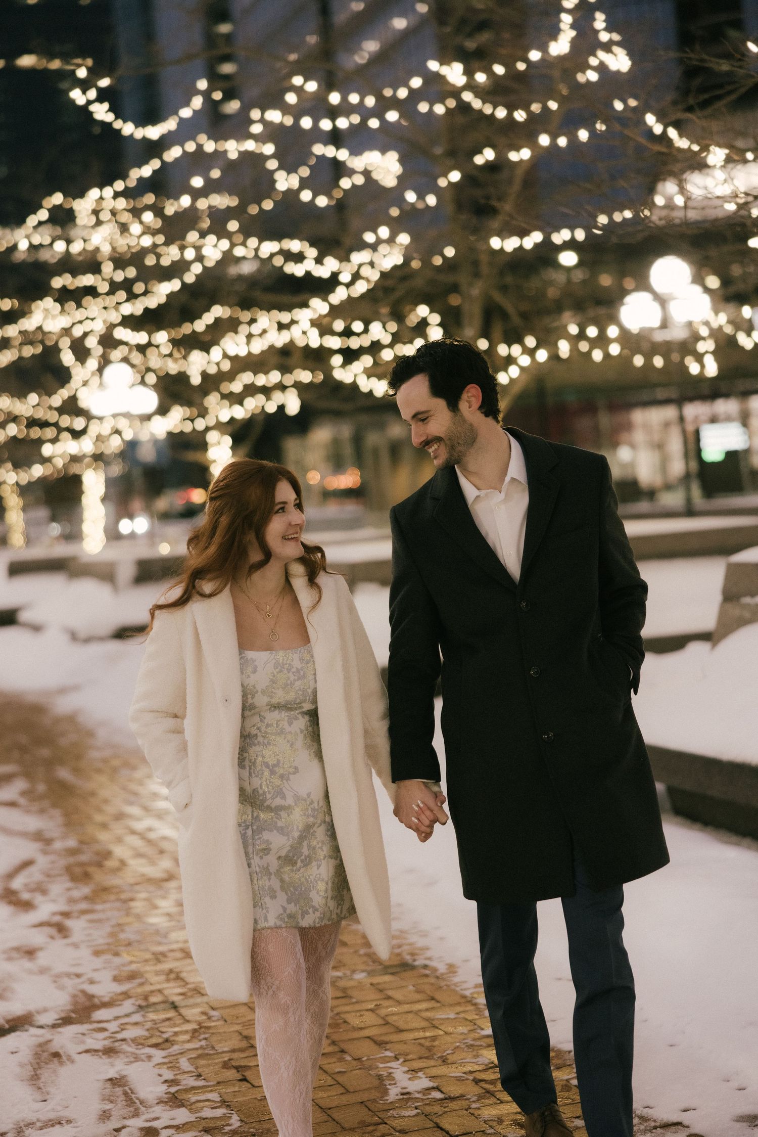 A couple walks hand in hand on a snowy evening beneath twinkling string lights in a romantic winter setting.