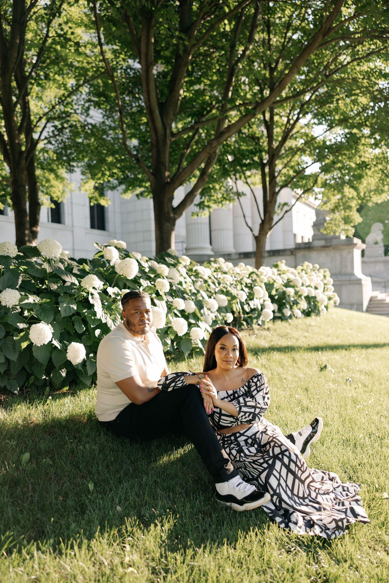 Couple sitting together on a grassy slope under shady trees, sharing a relaxing summer moment in white and black outfits.