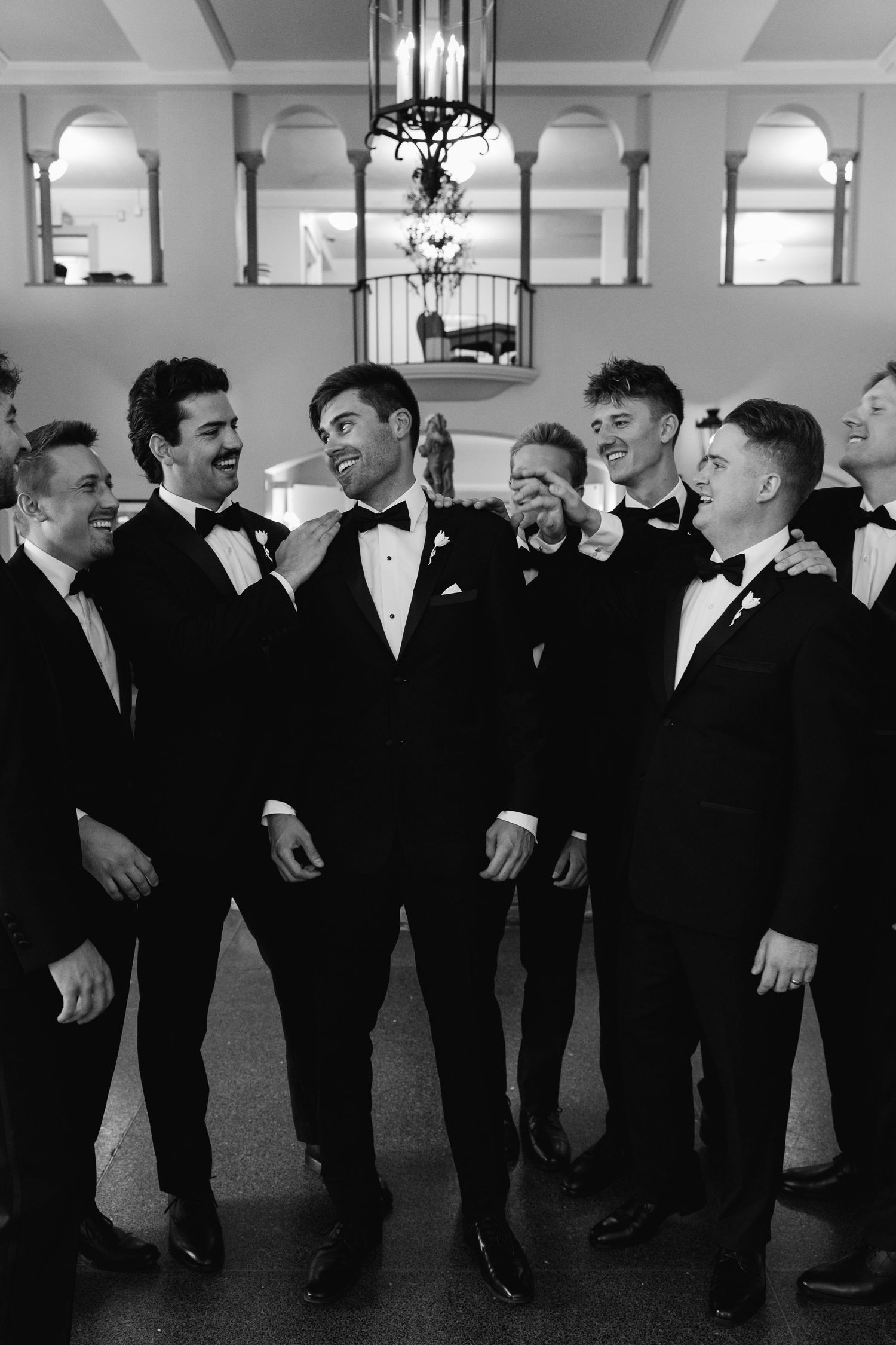 Groom and groomsmen laughing together in black tuxedos and bow ties inside an elegant venue.