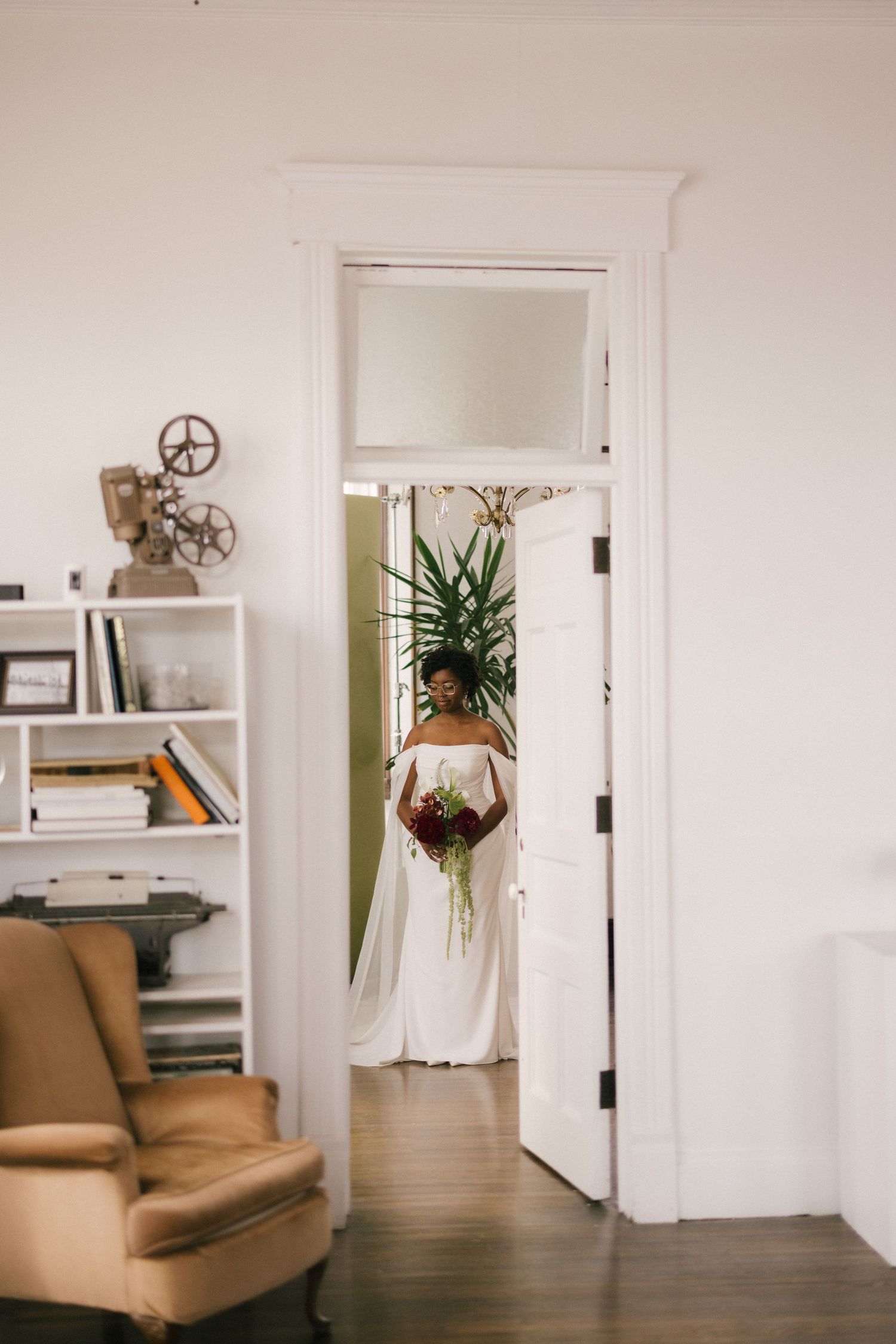 Bride in off-shoulder white gown holding bouquet stands in doorway of elegant room with bookshelf and armchair.