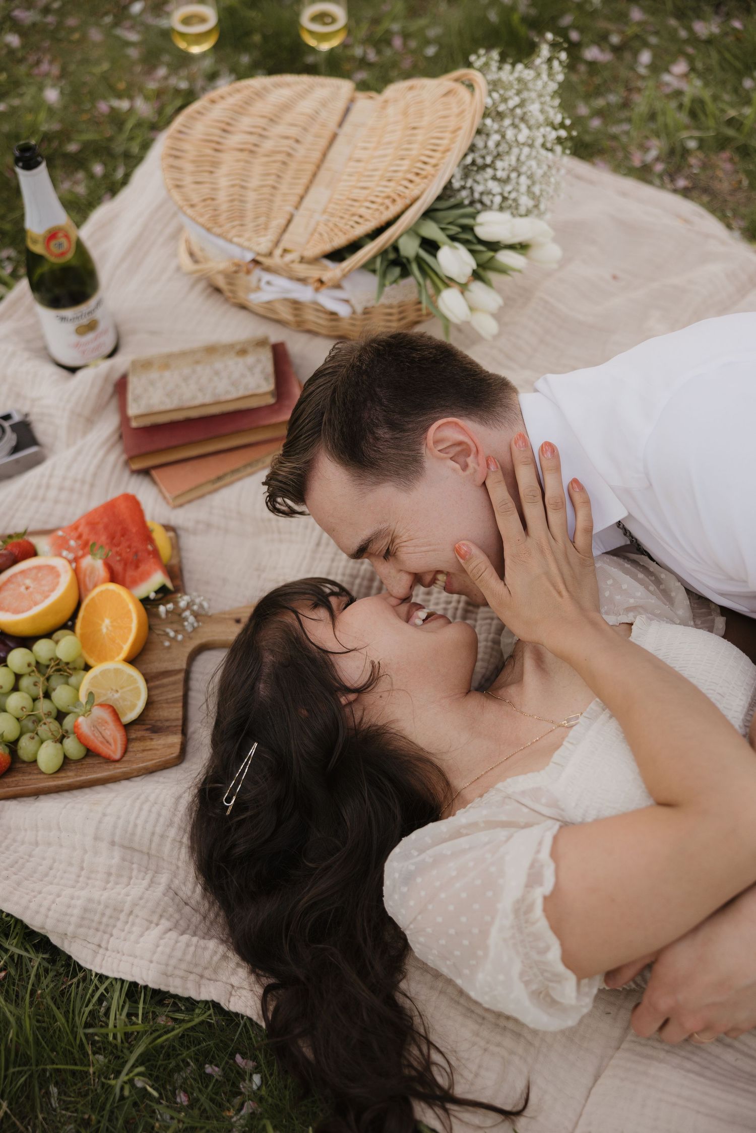 Romantic picnic setup with fruit platter, wine, wicker basket and couple sharing intimate moment on white blanket outdoors.