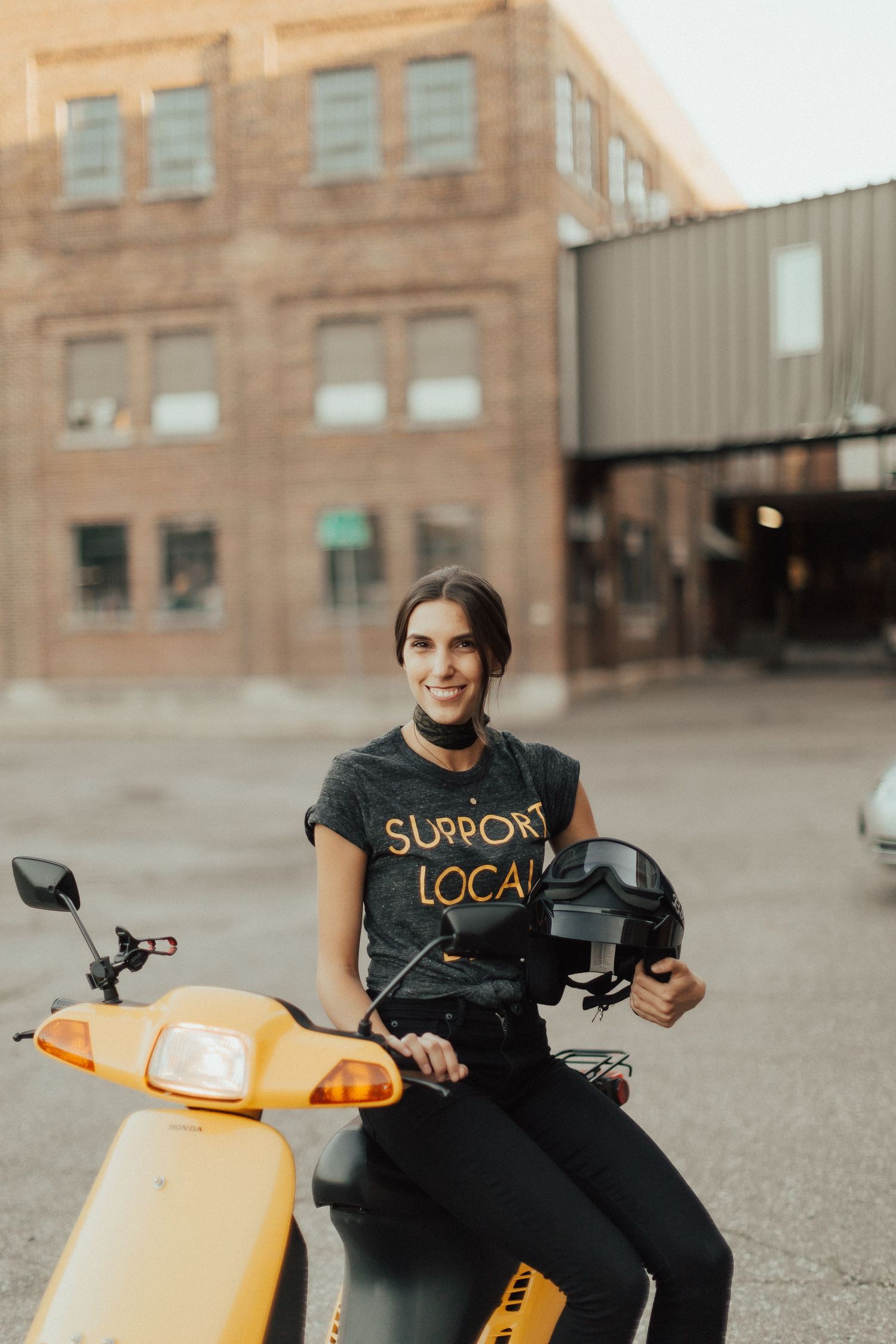 Person on yellow scooter wearing Support Local t-shirt in urban setting.