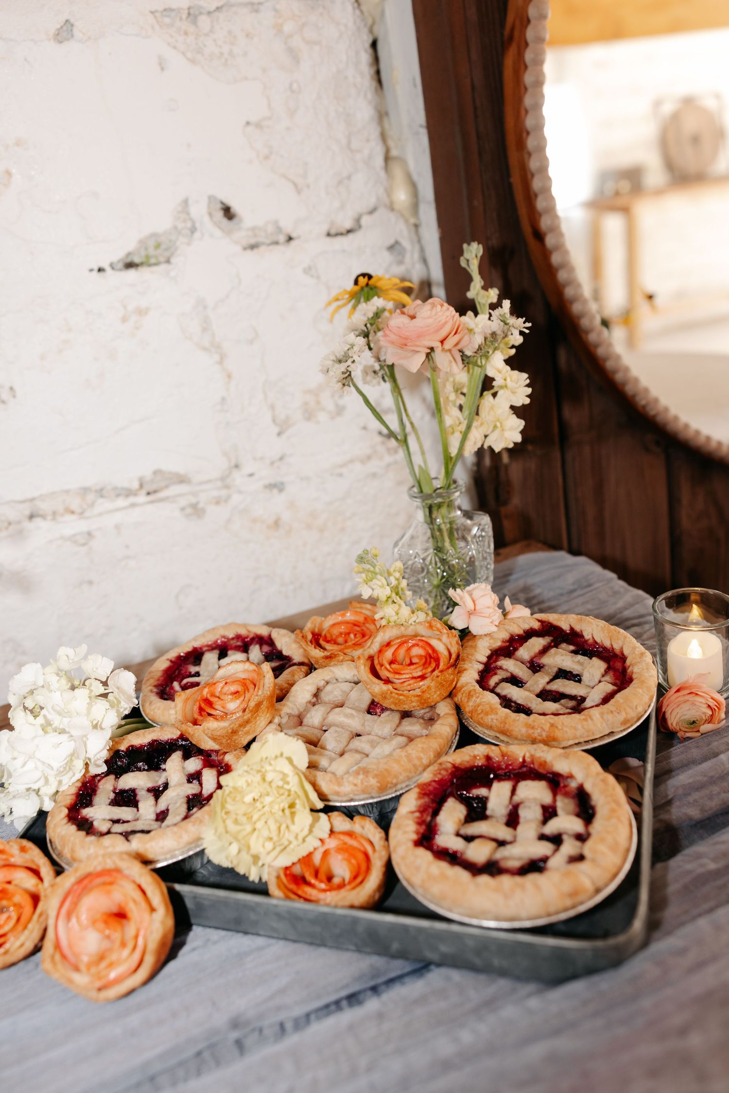 An elegant rustic dessert display featuring fresh pies decorated with lattice tops and coral roses on a gray tablecloth.