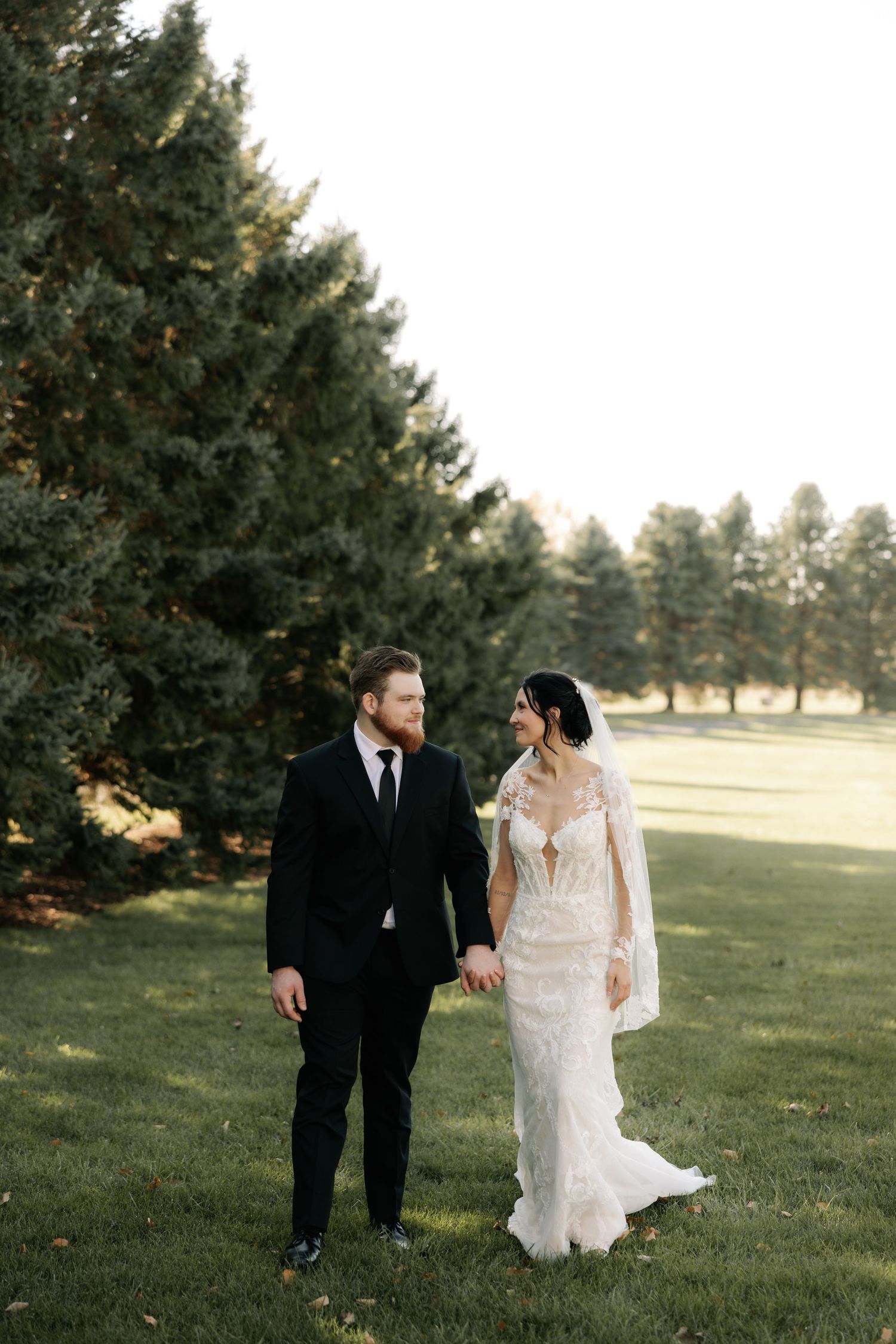 A couple in wedding attire walks together on a grassy field with evergreen trees in the background during golden hour.