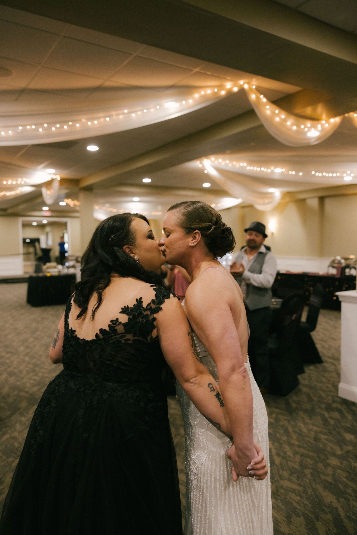 Two people share an intimate kiss during a wedding reception in a decorated ballroom with string lights.