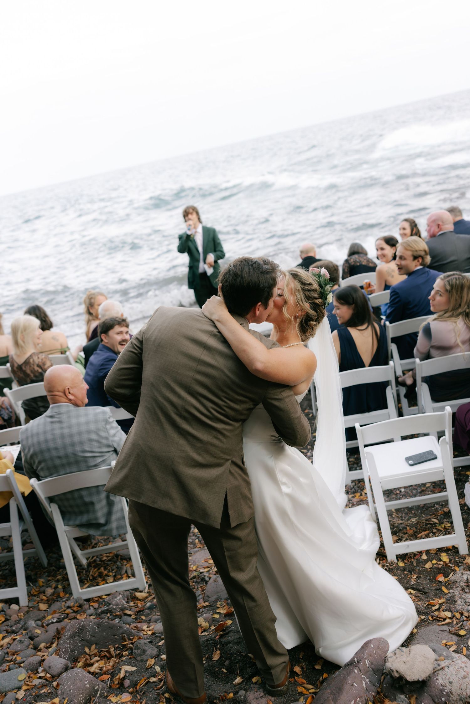 Wedding ceremony guests seated in white chairs on a rocky beach overlooking the ocean.