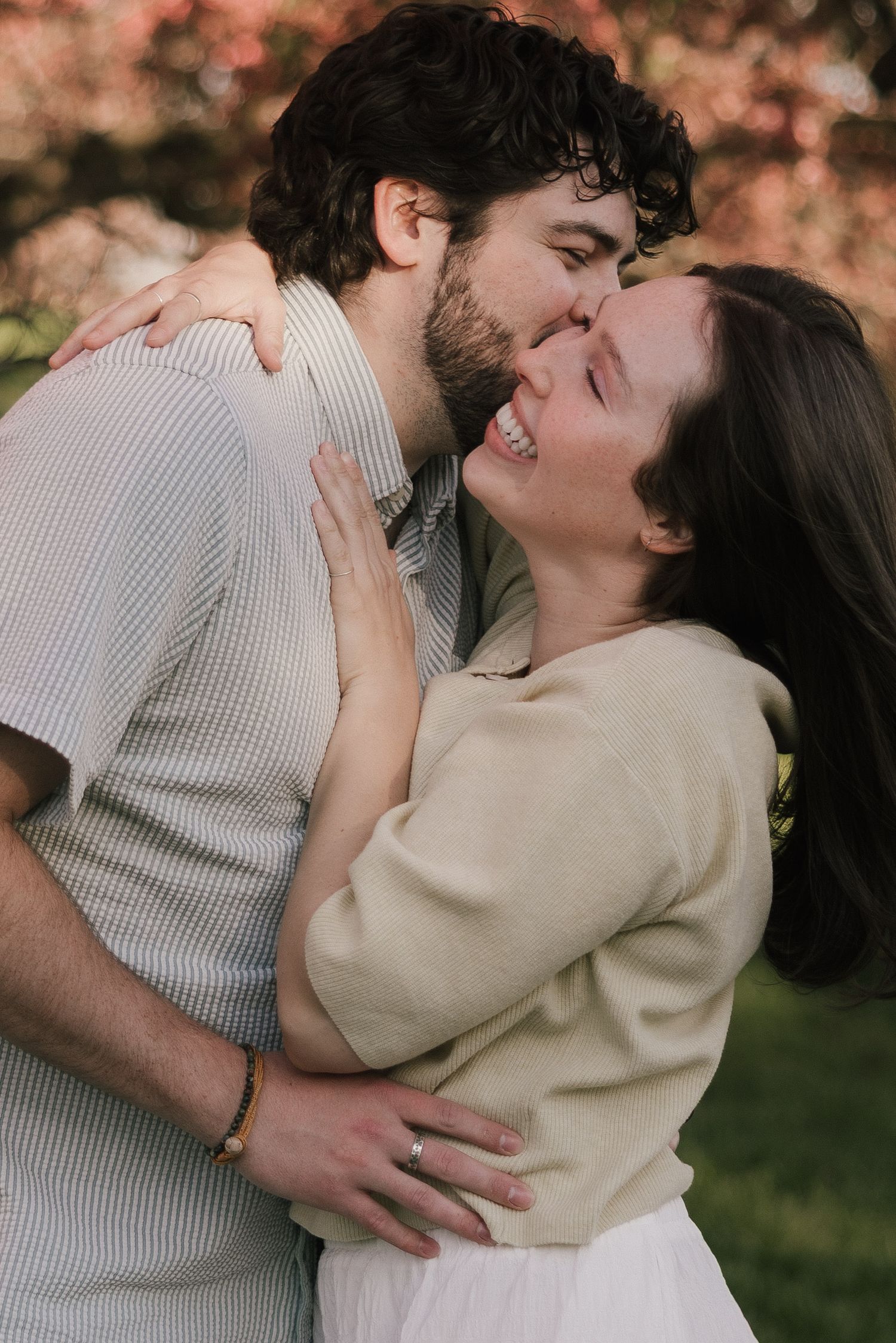 A happy couple embraces and shares a kiss outdoors, surrounded by pink blossoming trees in soft natural light.