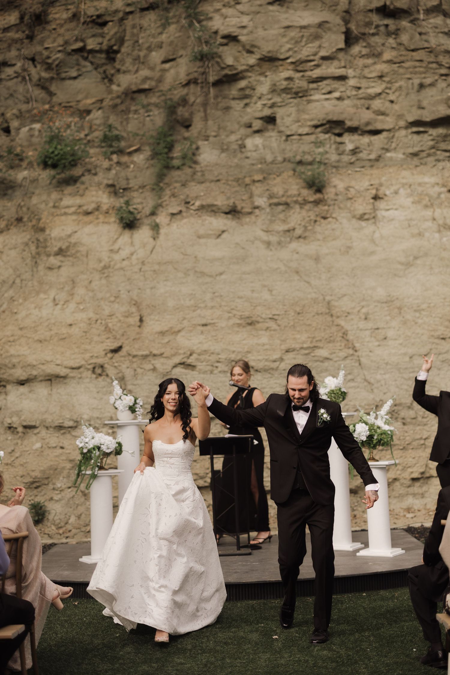 Bride and groom celebrate after wedding ceremony outdoors against a dramatic rock cliff backdrop with floral arrangements.