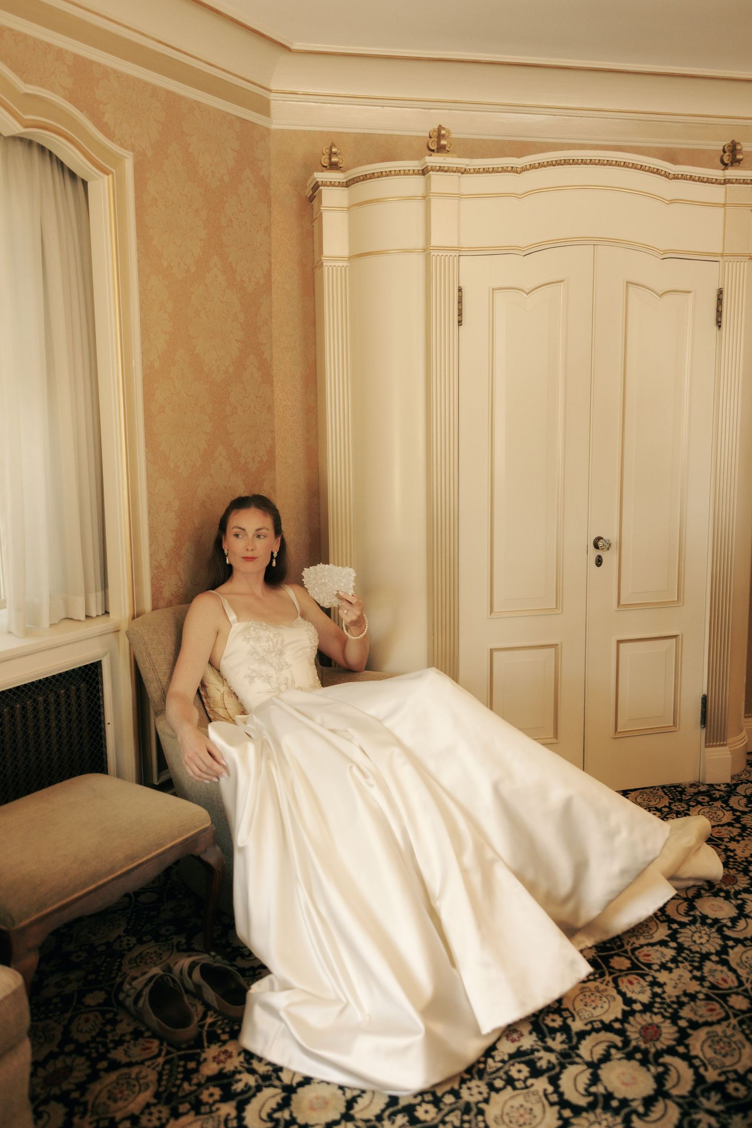 Bride in white satin gown relaxes on chair holding bouquet in elegant room with ornate wallpaper and classic wardrobe.