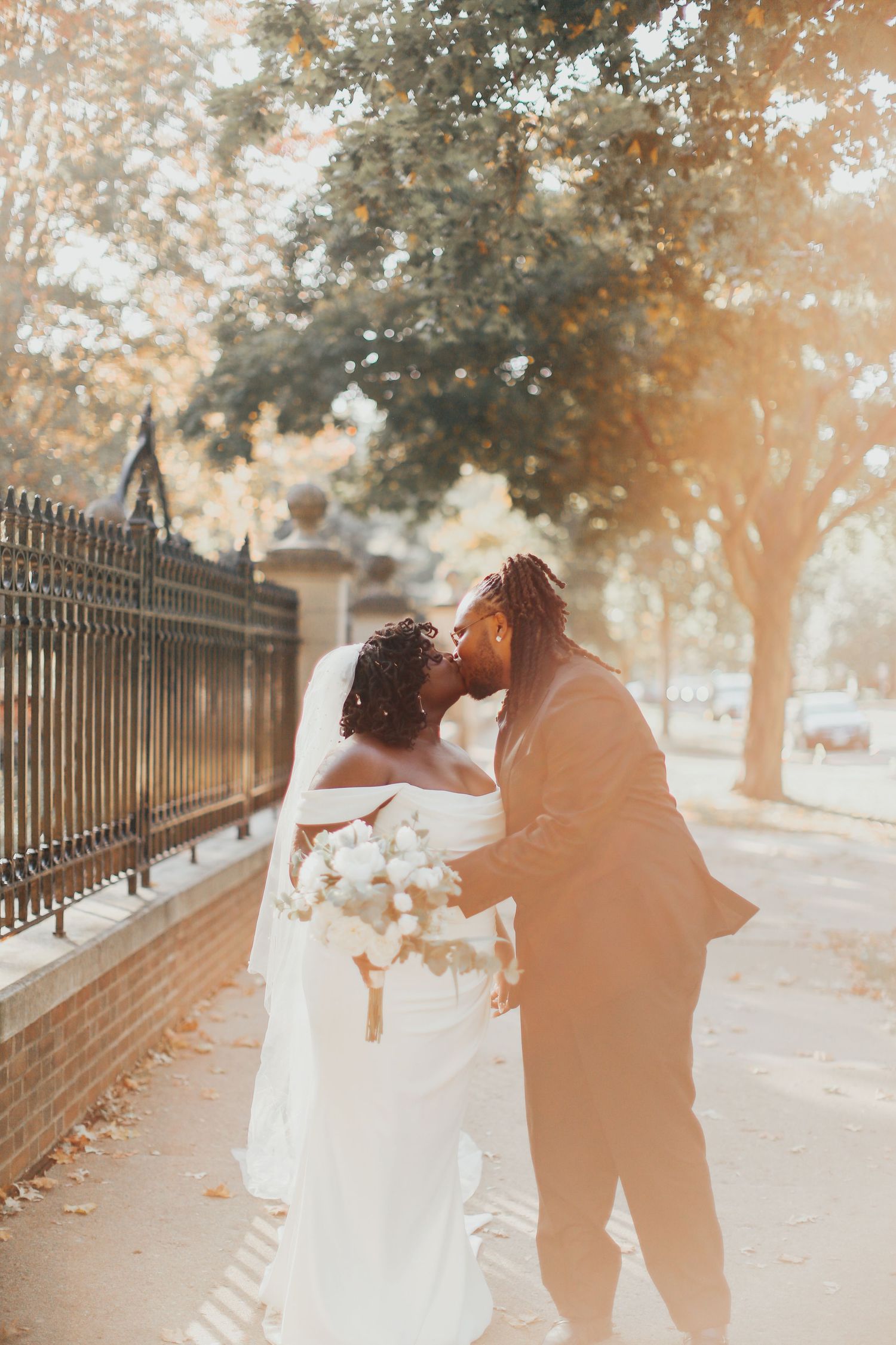 Romantic sunset portrait of newlyweds embracing by iron fence and brick wall.