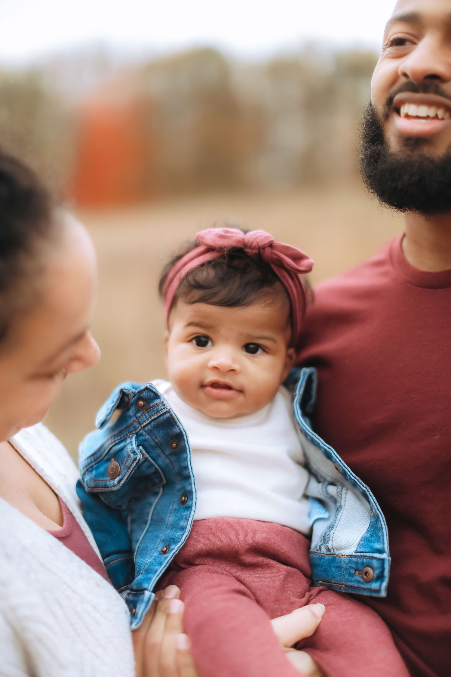 Baby in denim jacket and pink pants held lovingly by parents outdoors.
