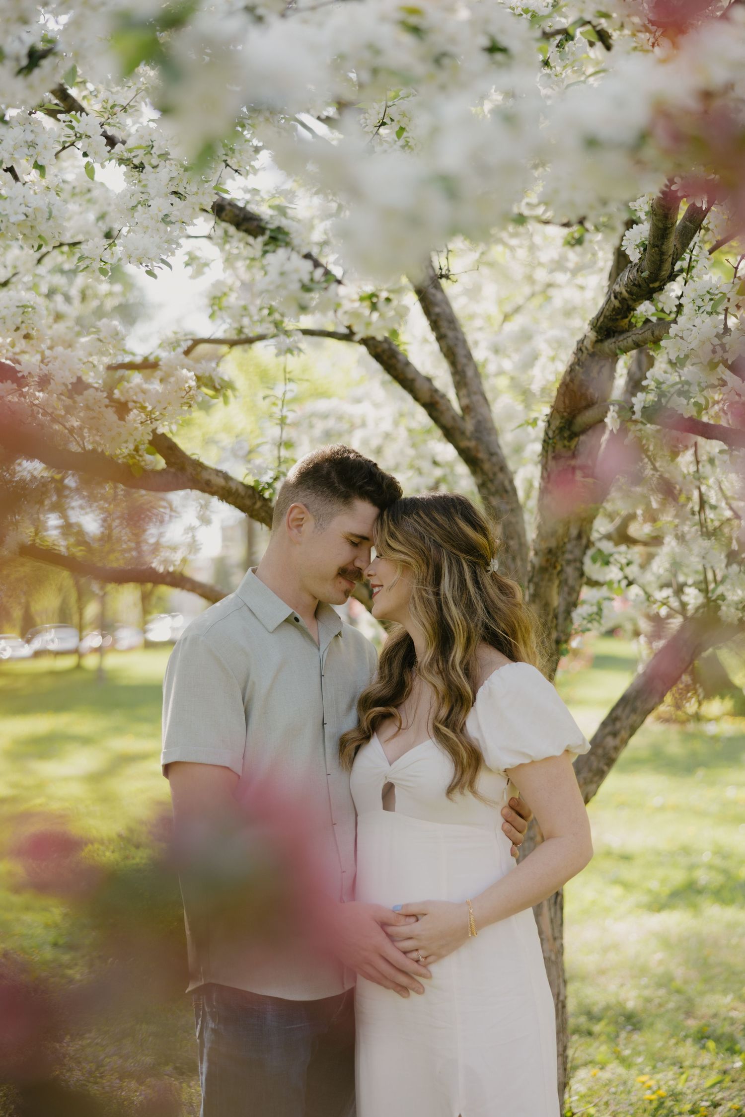A couple shares an intimate moment under blooming white spring tree blossoms in a sunlit garden setting.