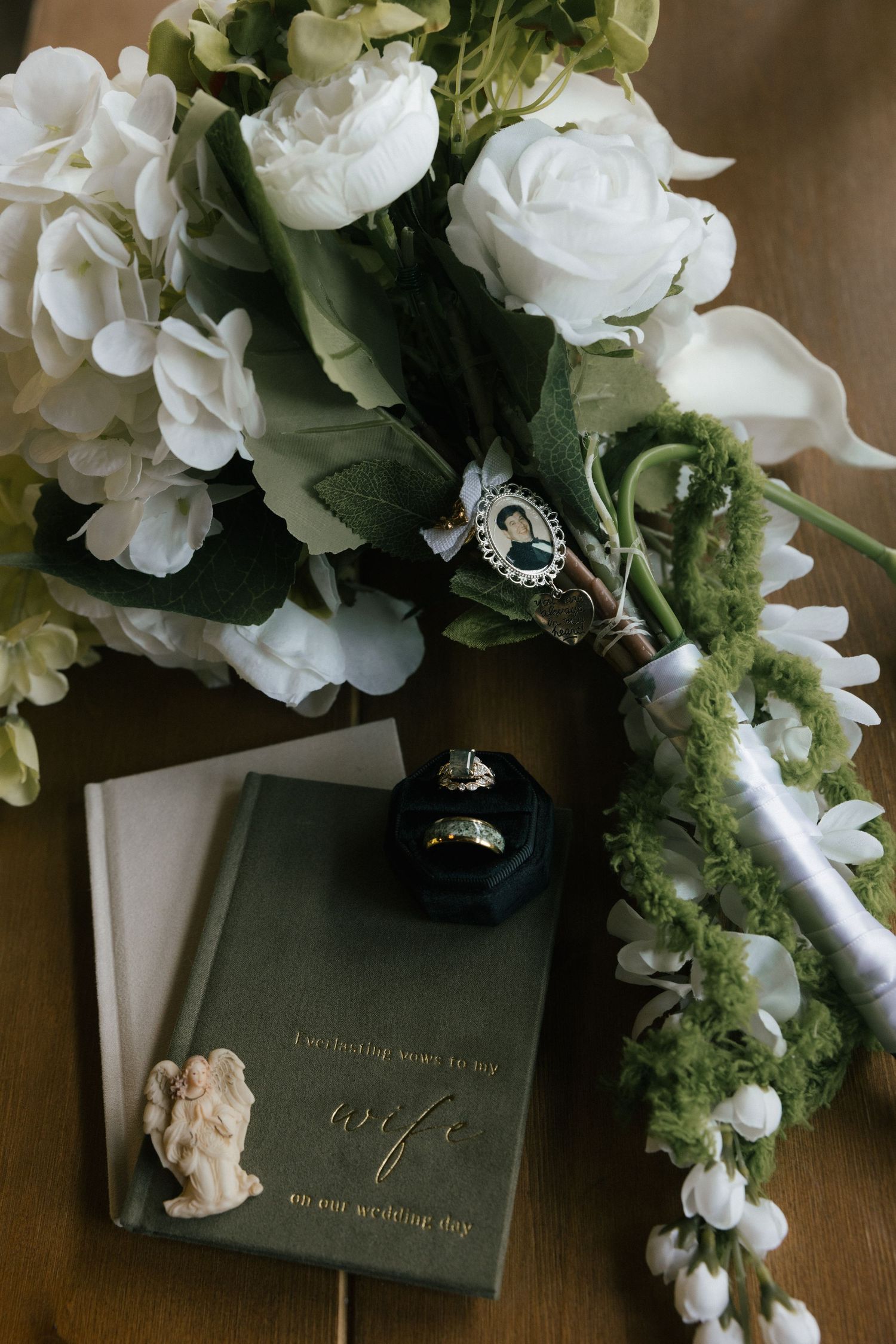 Elegant wedding flat lay with white floral bouquet, ring box, gold bands, angel figurine, and a wife vow book on wood.