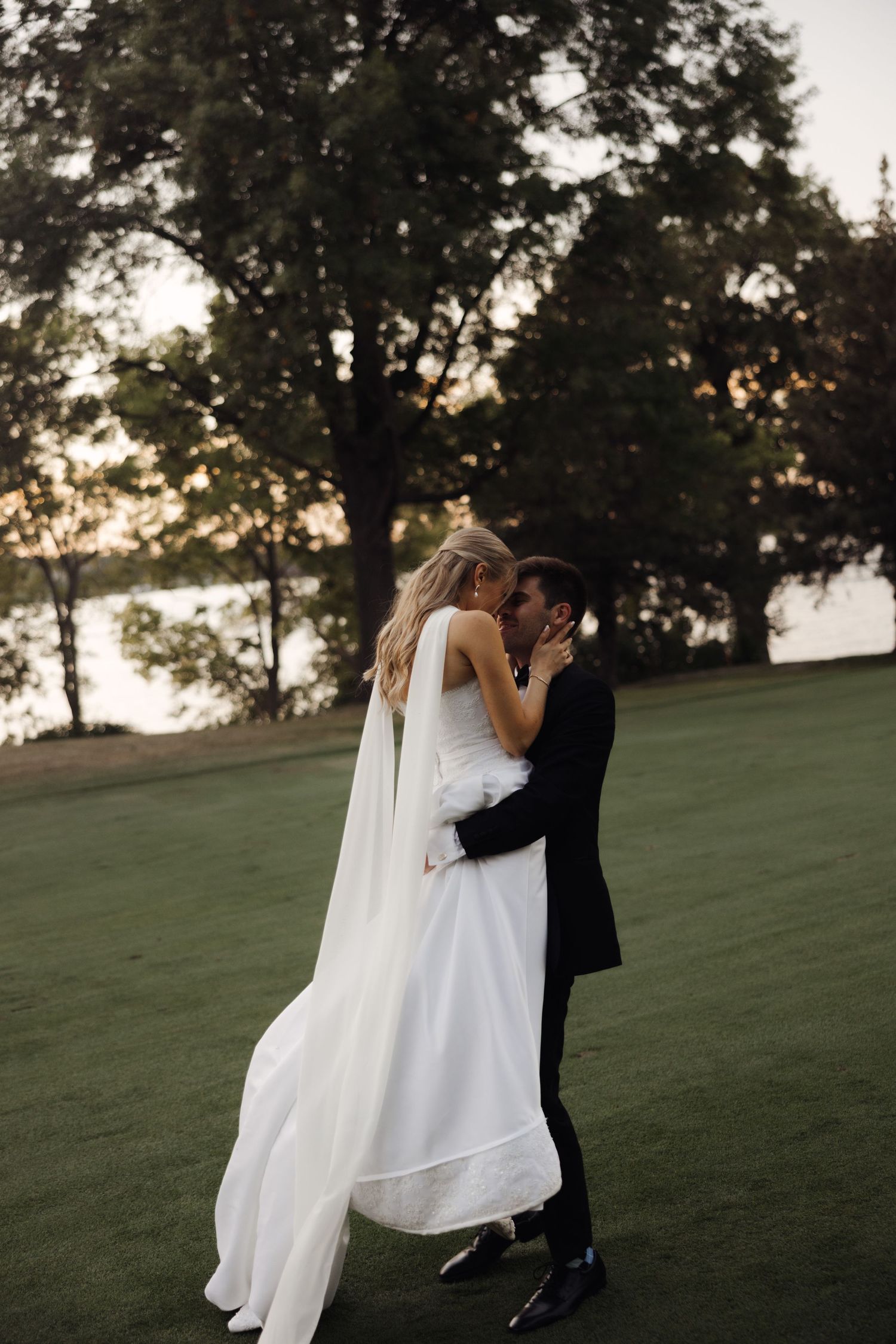 Bride and groom embracing on green lawn at sunset with trees and water in background at outdoor wedding.
