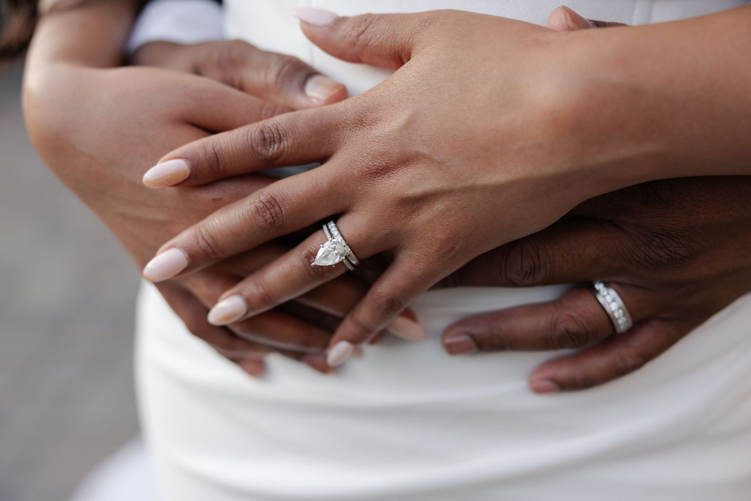 Two hands wearing wedding rings rest gently against white fabric, showcasing an elegant diamond engagement ring.