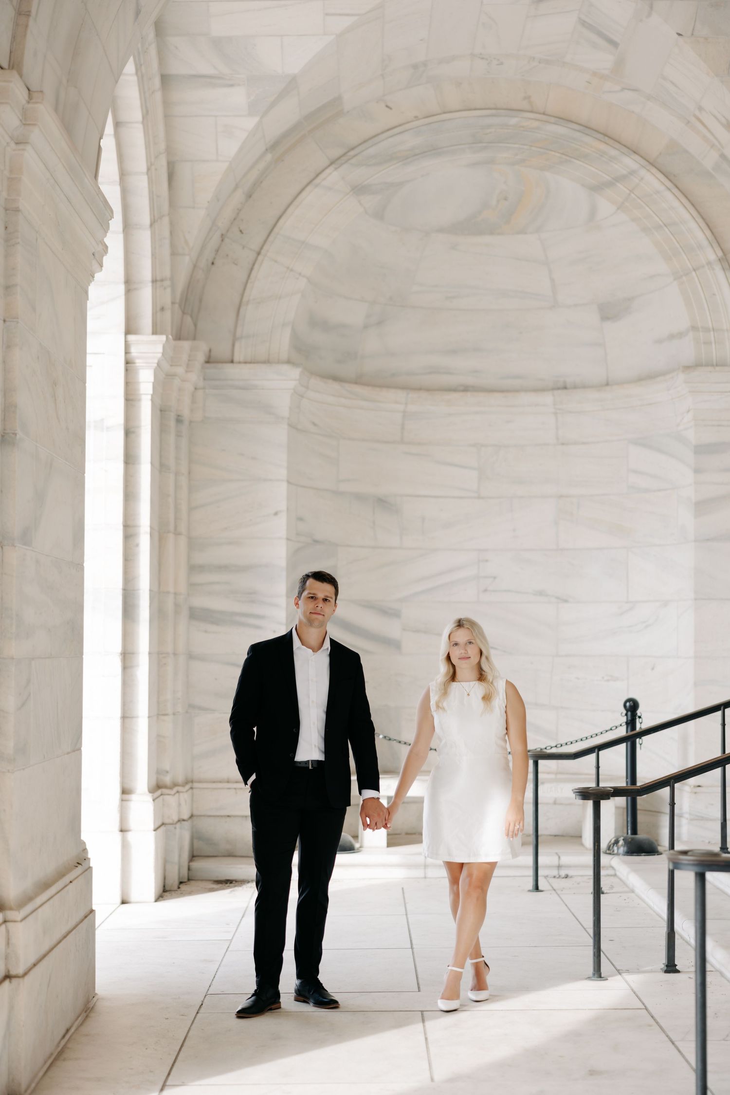 A couple poses in a bright white marble corridor with arched ceilings and black railings.