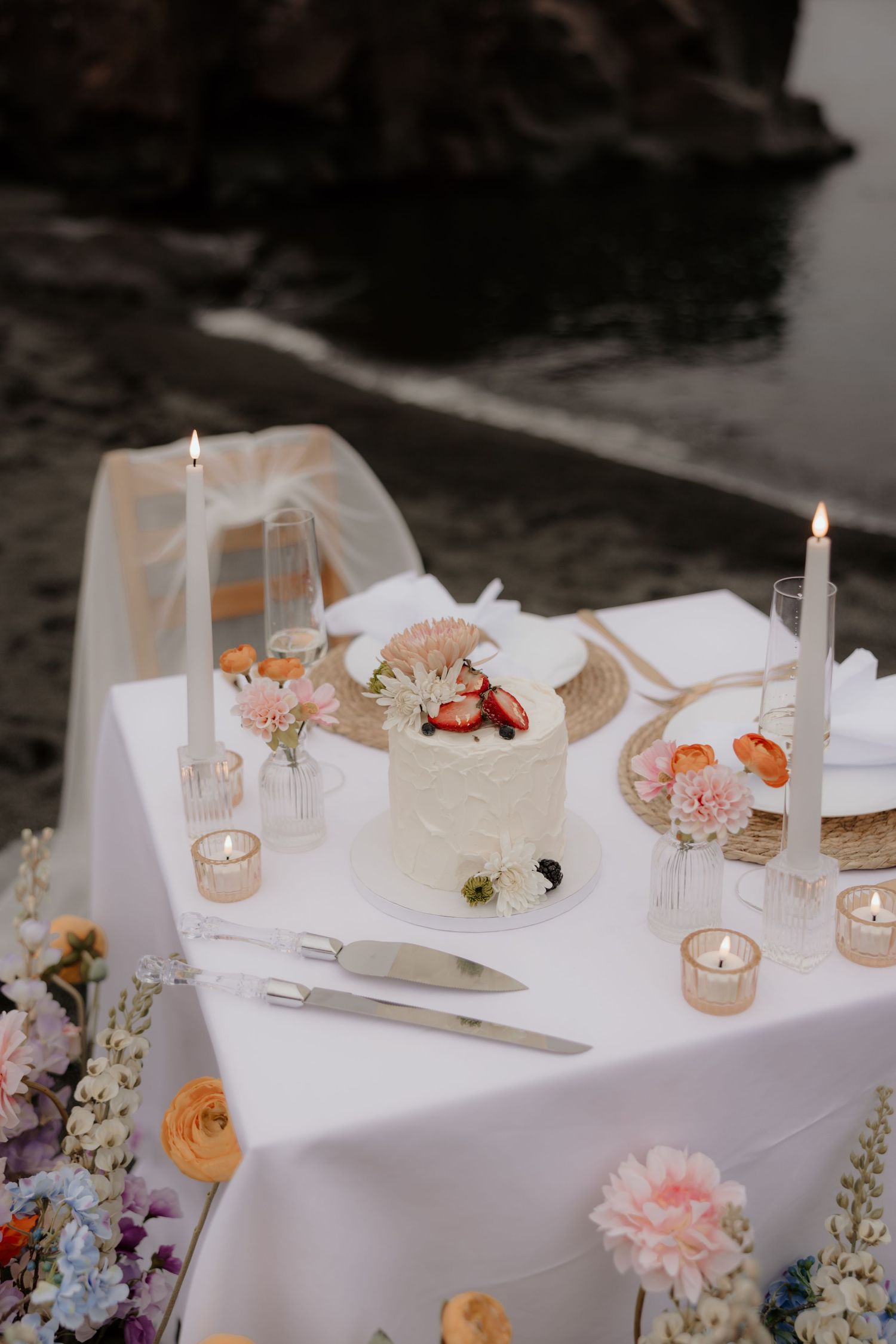 Romantic table setting with white tablecloth, lit candles, floral arrangements and a small tiered cake on coastal background.