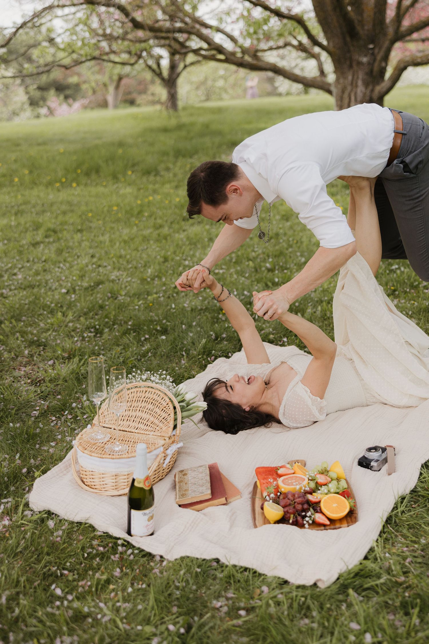 A couple enjoys a romantic outdoor picnic on a white blanket with a wicker basket and snacks under large trees.