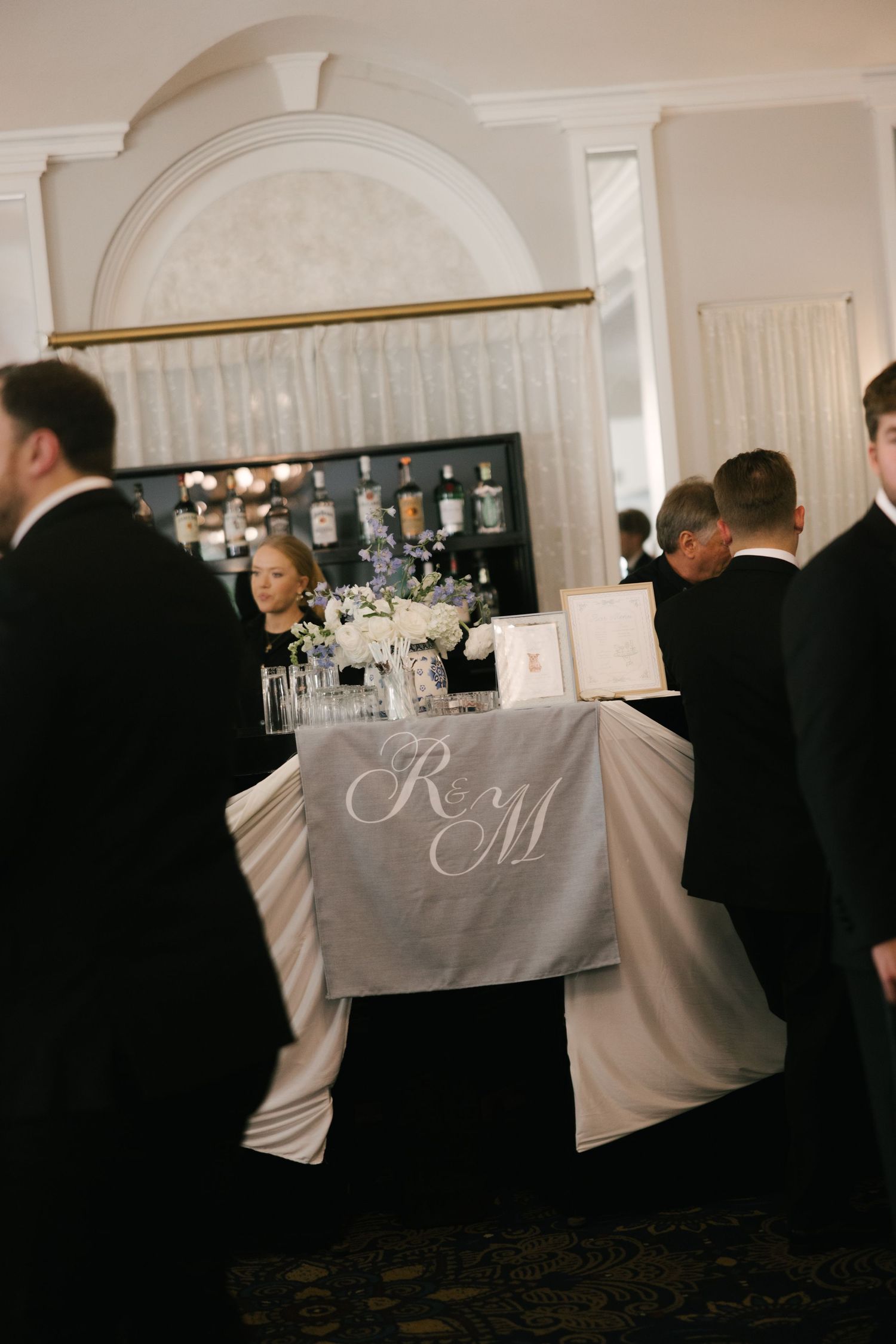 Elegant wedding sweetheart table with monogrammed R&M linen, white floral centerpiece, and guests in formal black tie attire.
