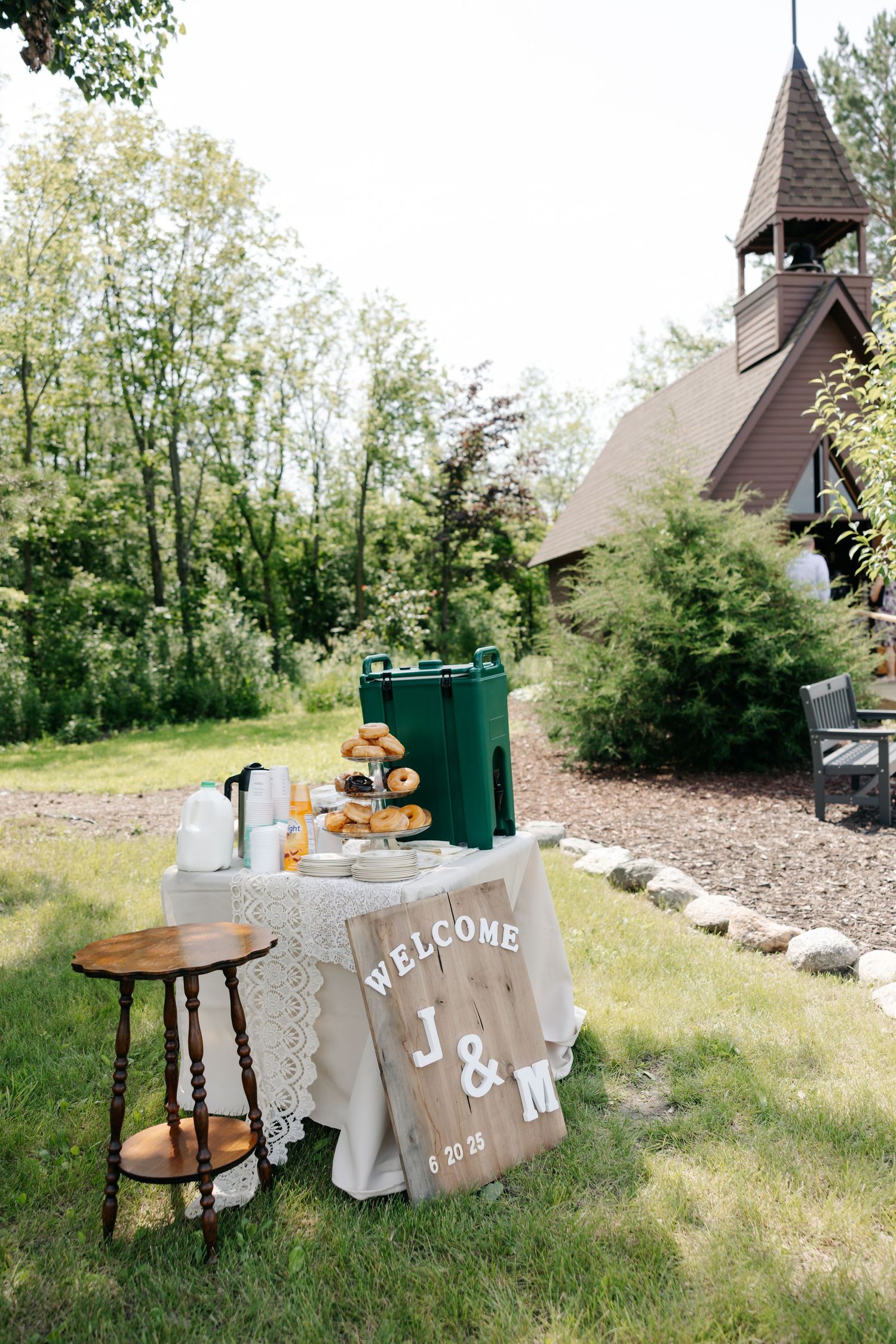 Wedding decor featuring rustic wooden furniture and welcome signs displayed on a grassy hillside with a chapel in the background.