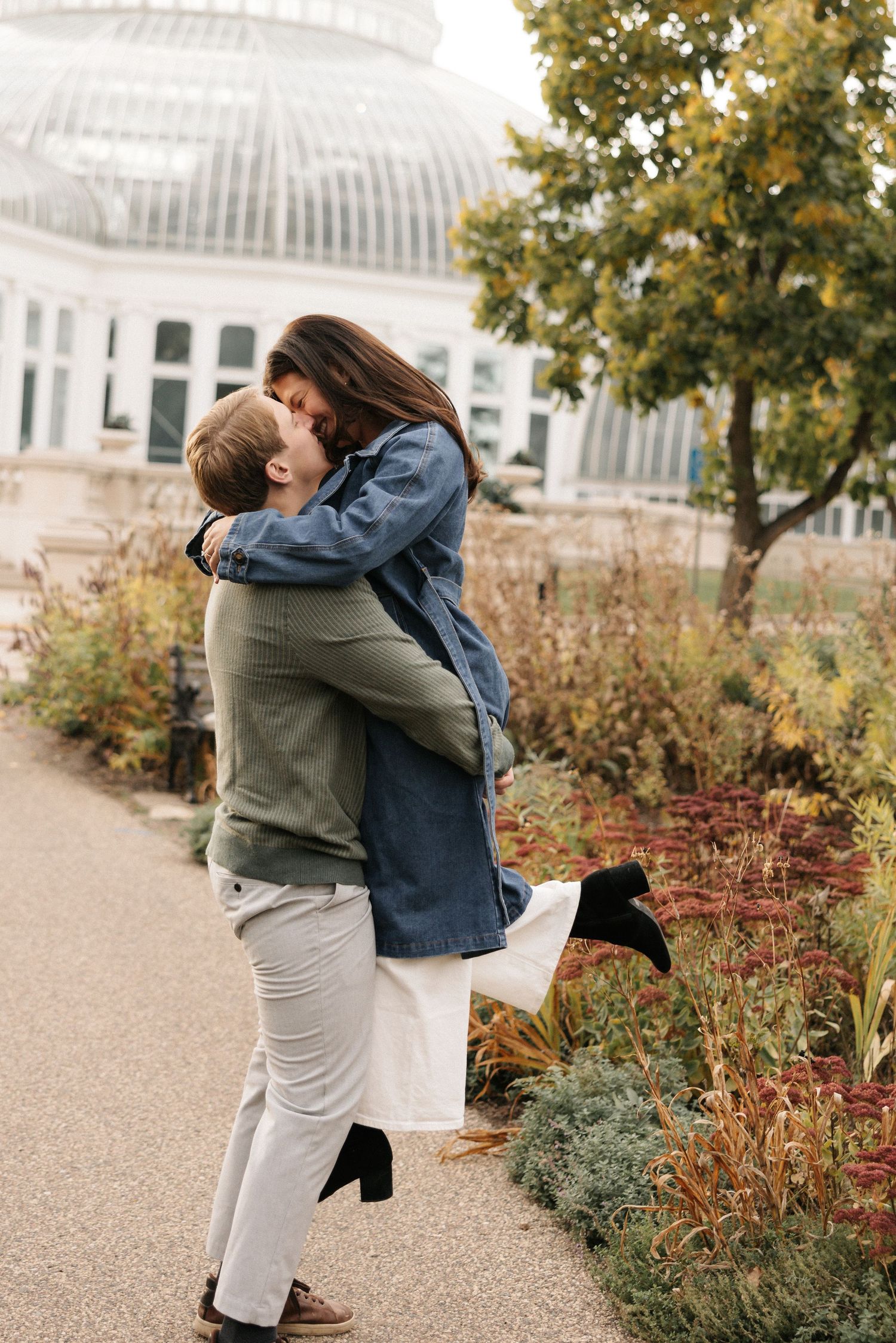 A couple embraces and lifts each other up on a garden path near a greenhouse on an autumn day.
