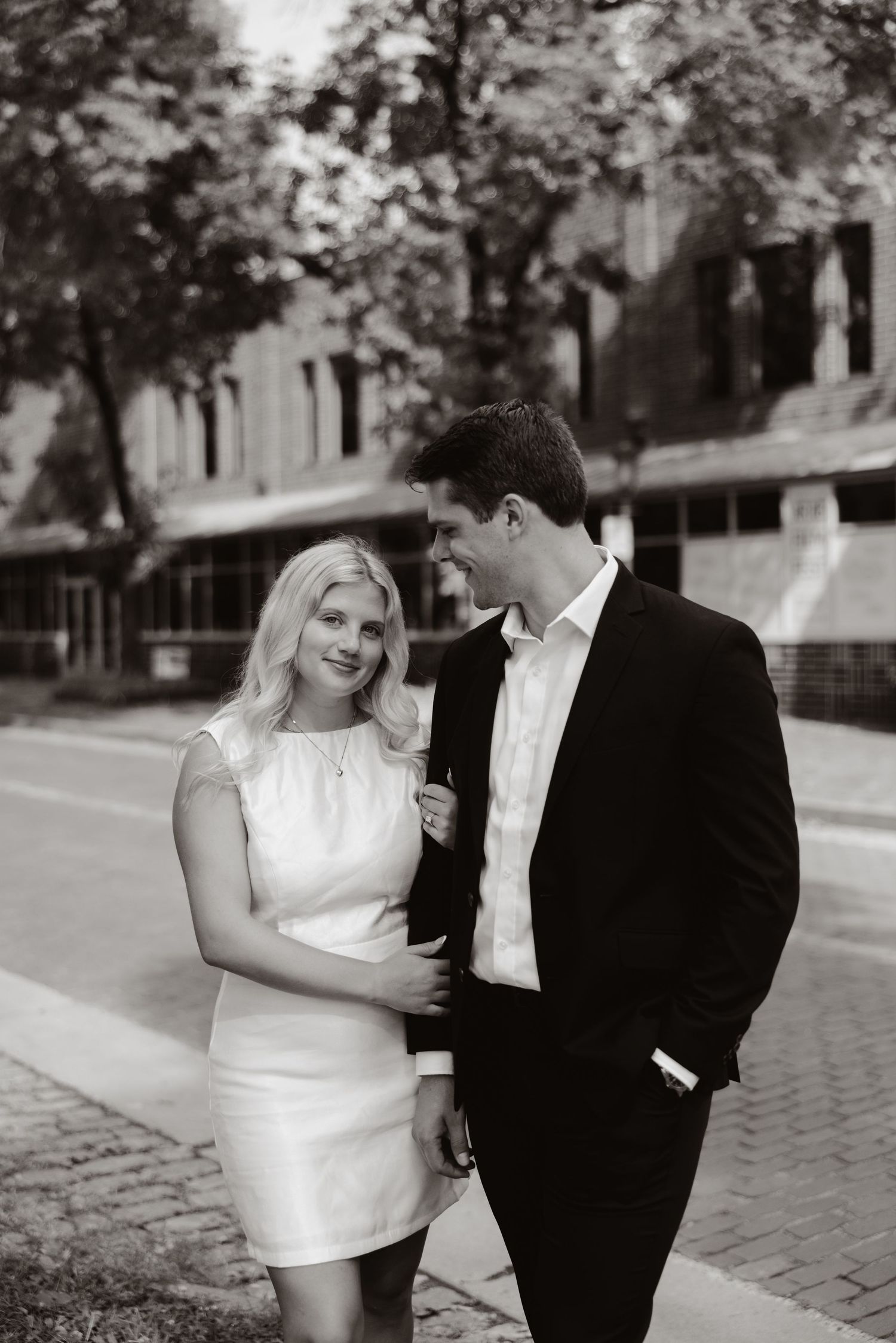 Black and white photo of a smiling couple in formal attire walking together on a city street.