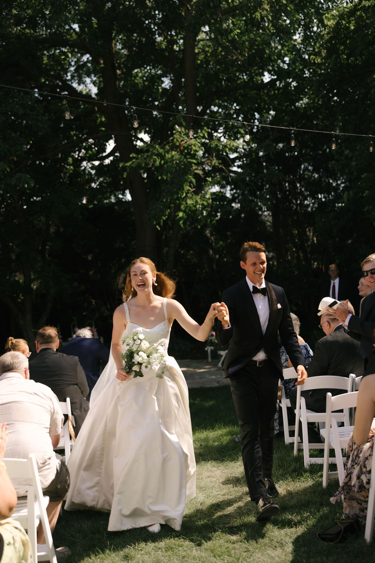 Joyful bride and groom walk down outdoor aisle after wedding ceremony, surrounded by guests seated in white chairs.