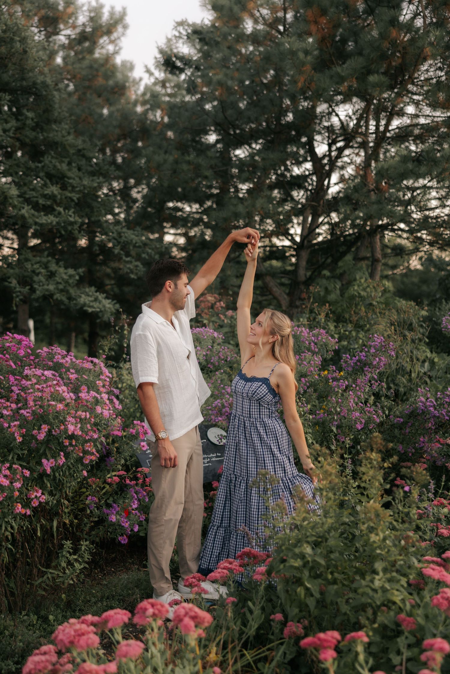 A couple shares a tender moment in a garden filled with vibrant pink flowers and lush greenery at sunset.