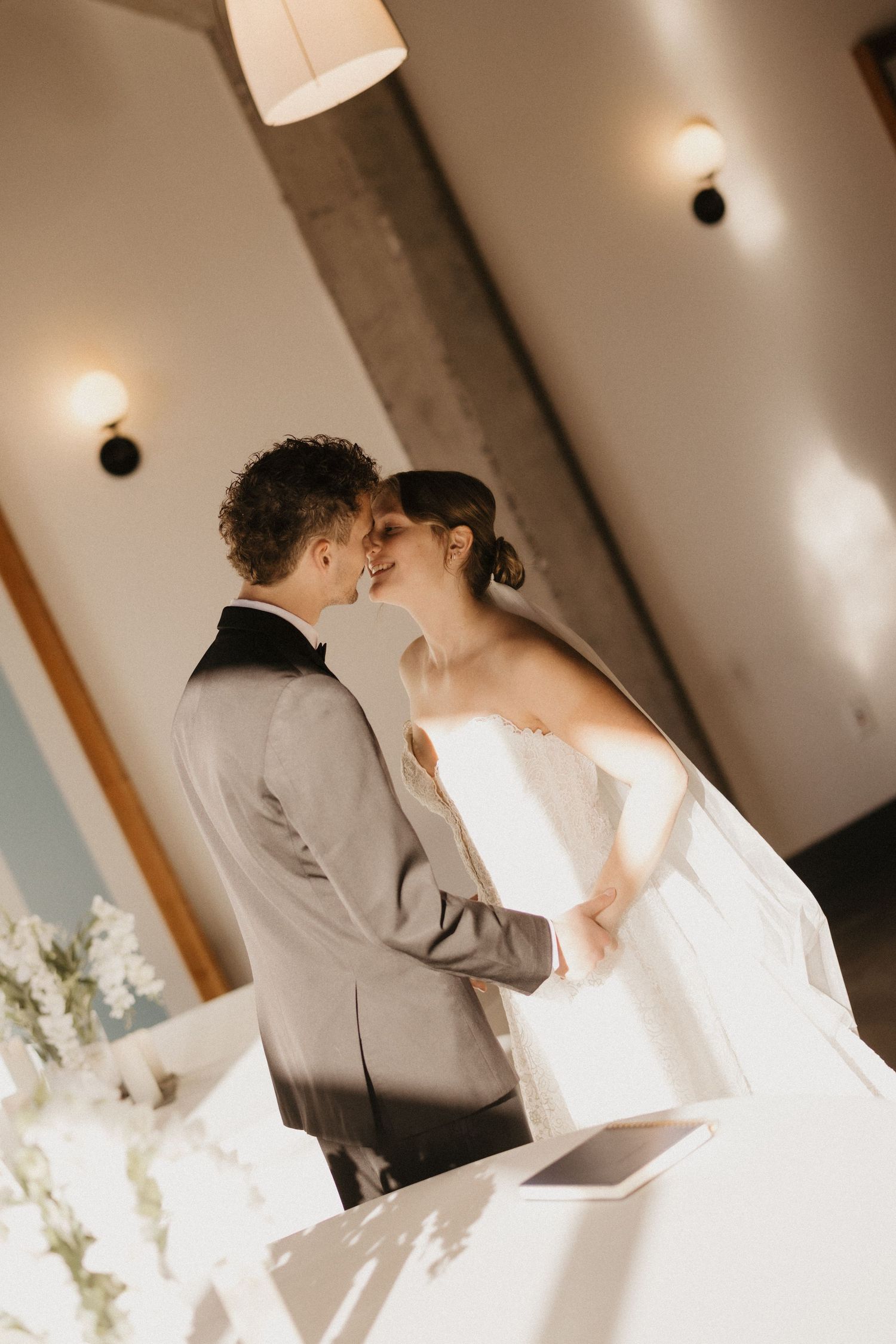 A couple in wedding attire share a tender moment in a sun-lit room with white decor and exposed wooden beams.