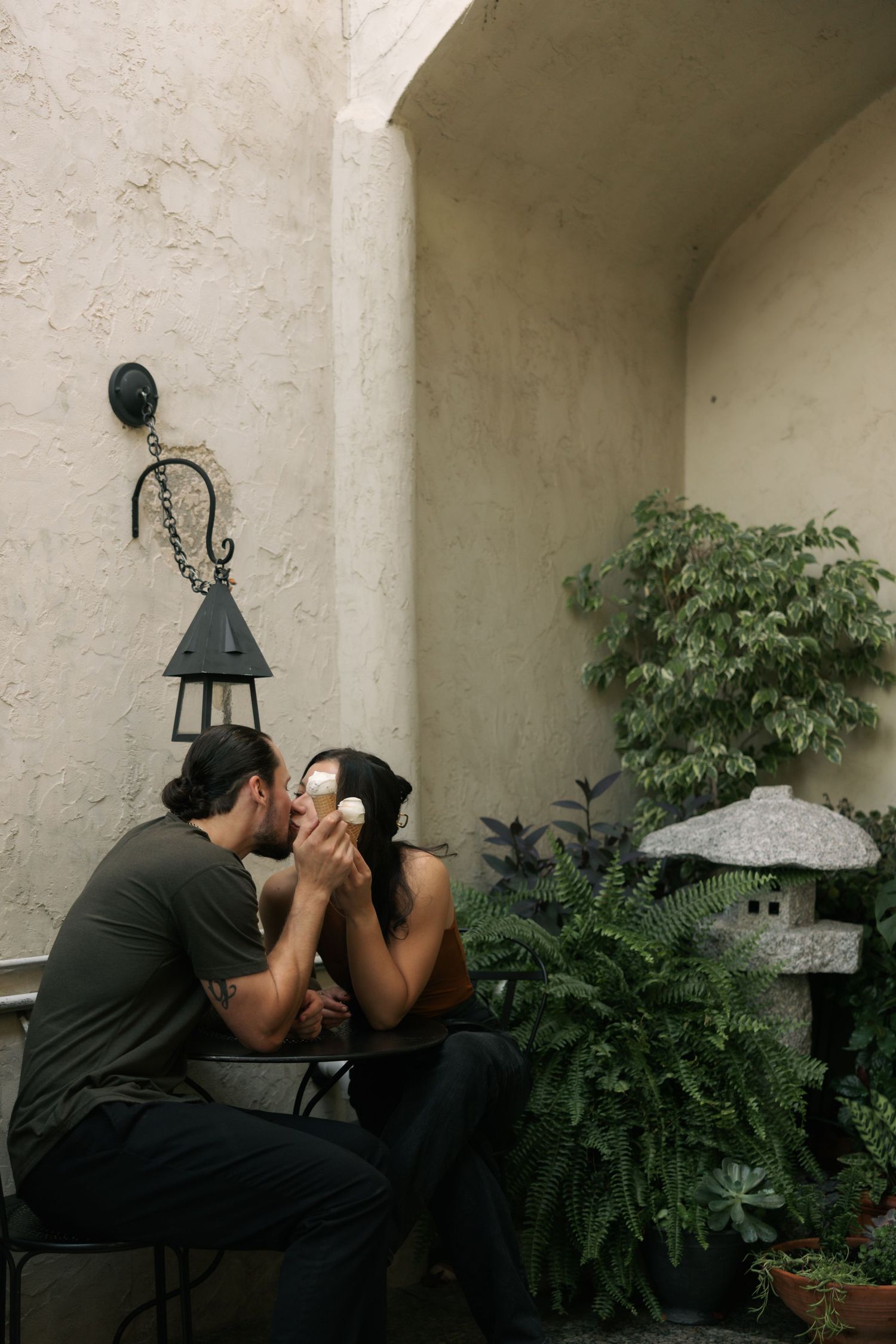 A couple shares an intimate moment on a patio bench surrounded by potted plants and white walls.