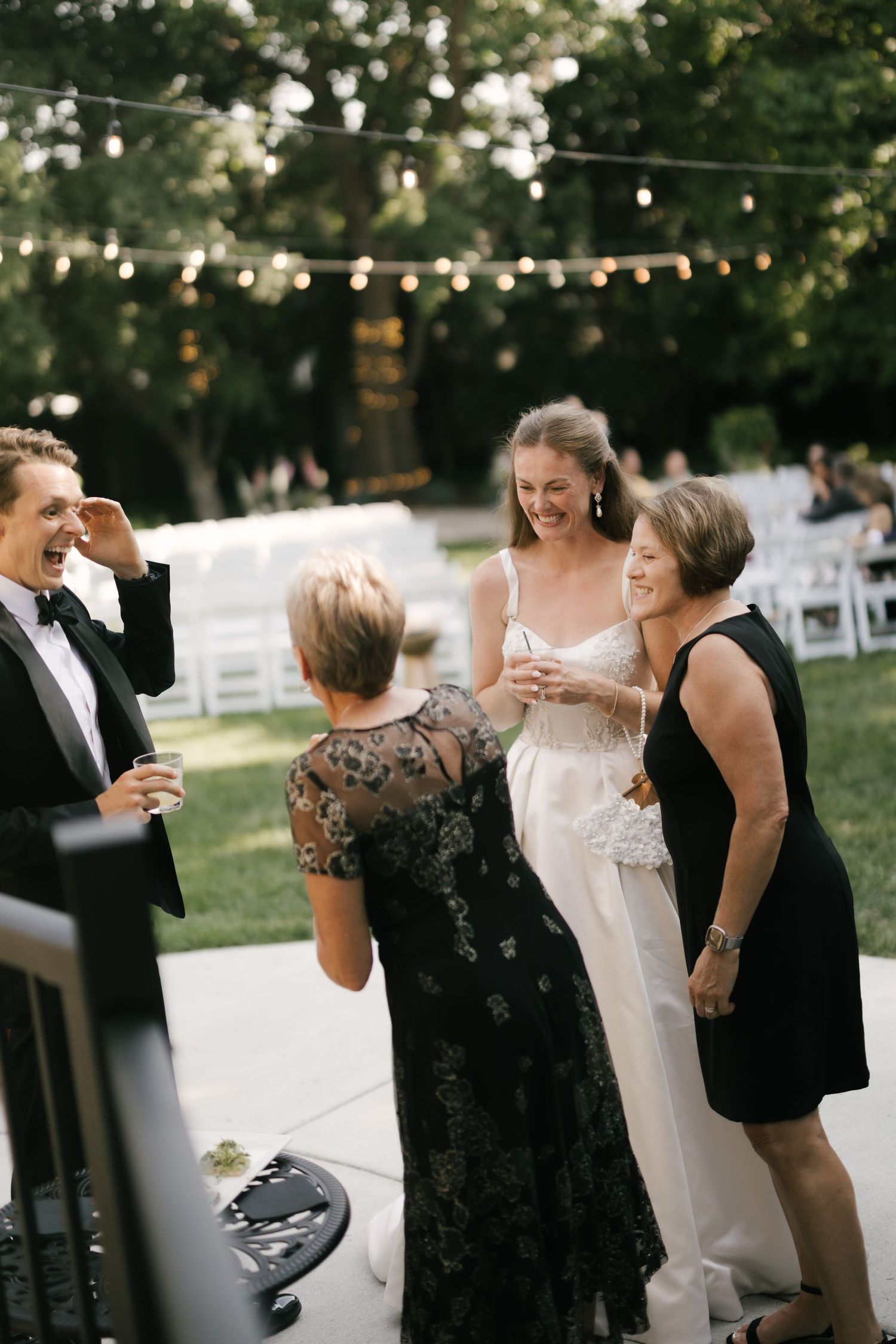 Bride in white gown laughing with guests at an outdoor wedding reception under string lights.