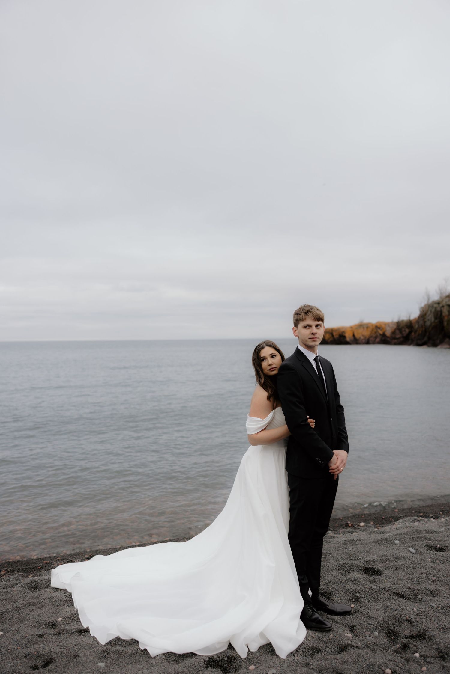 A couple poses back-to-back on a rocky beach with overcast skies and flowing white dress against dark waters.