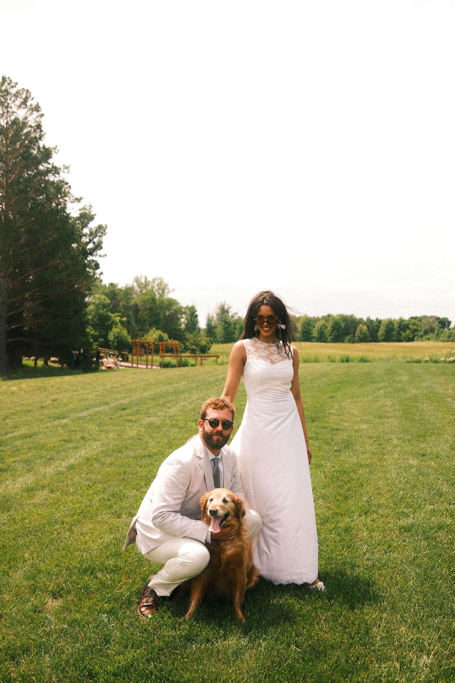 A couple in white outfits sitting together with their dog on a sunny grassy hill during a summer afternoon.