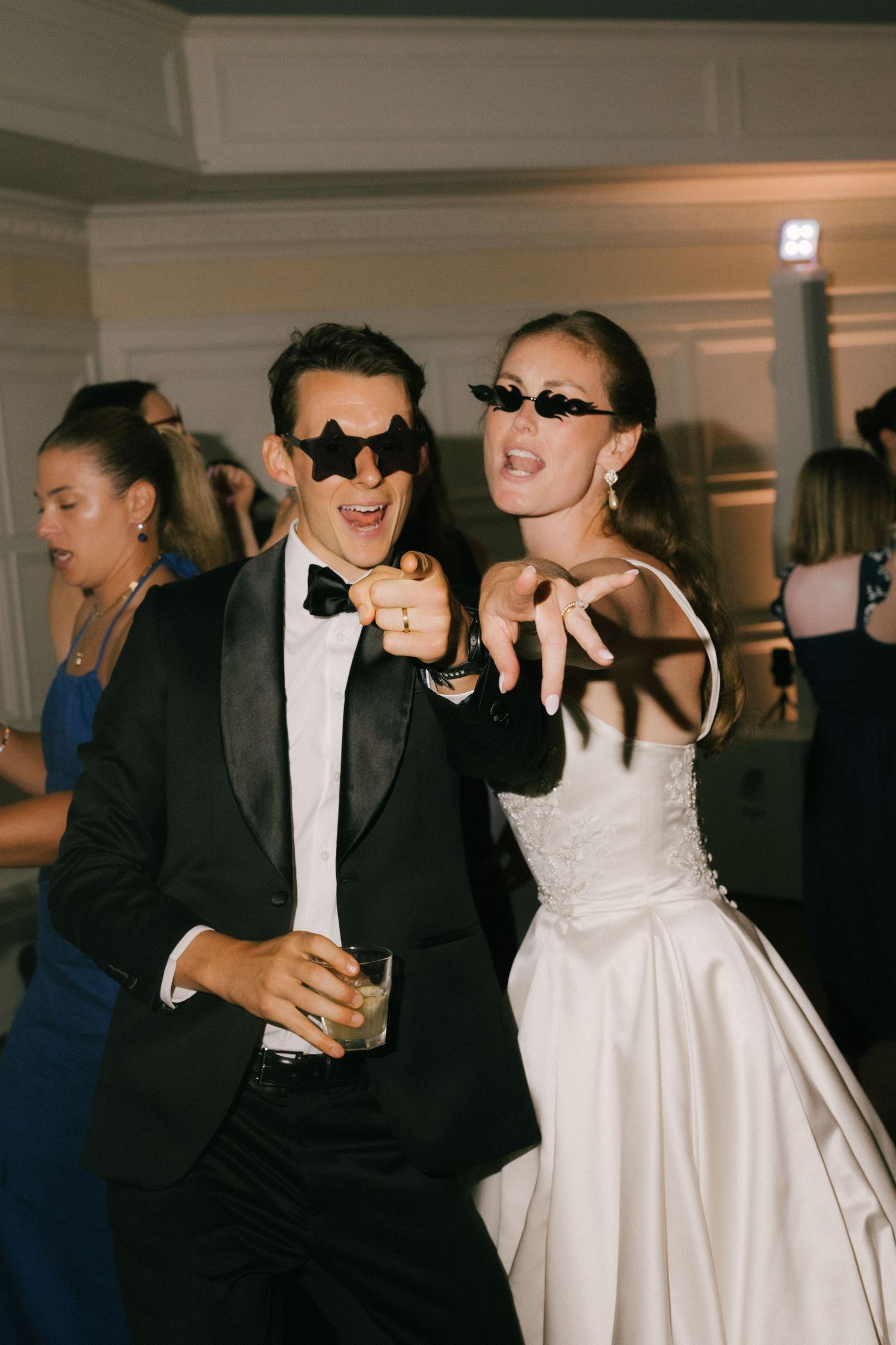 Bride and groom in sunglasses dancing and pointing at wedding reception in elegant venue.