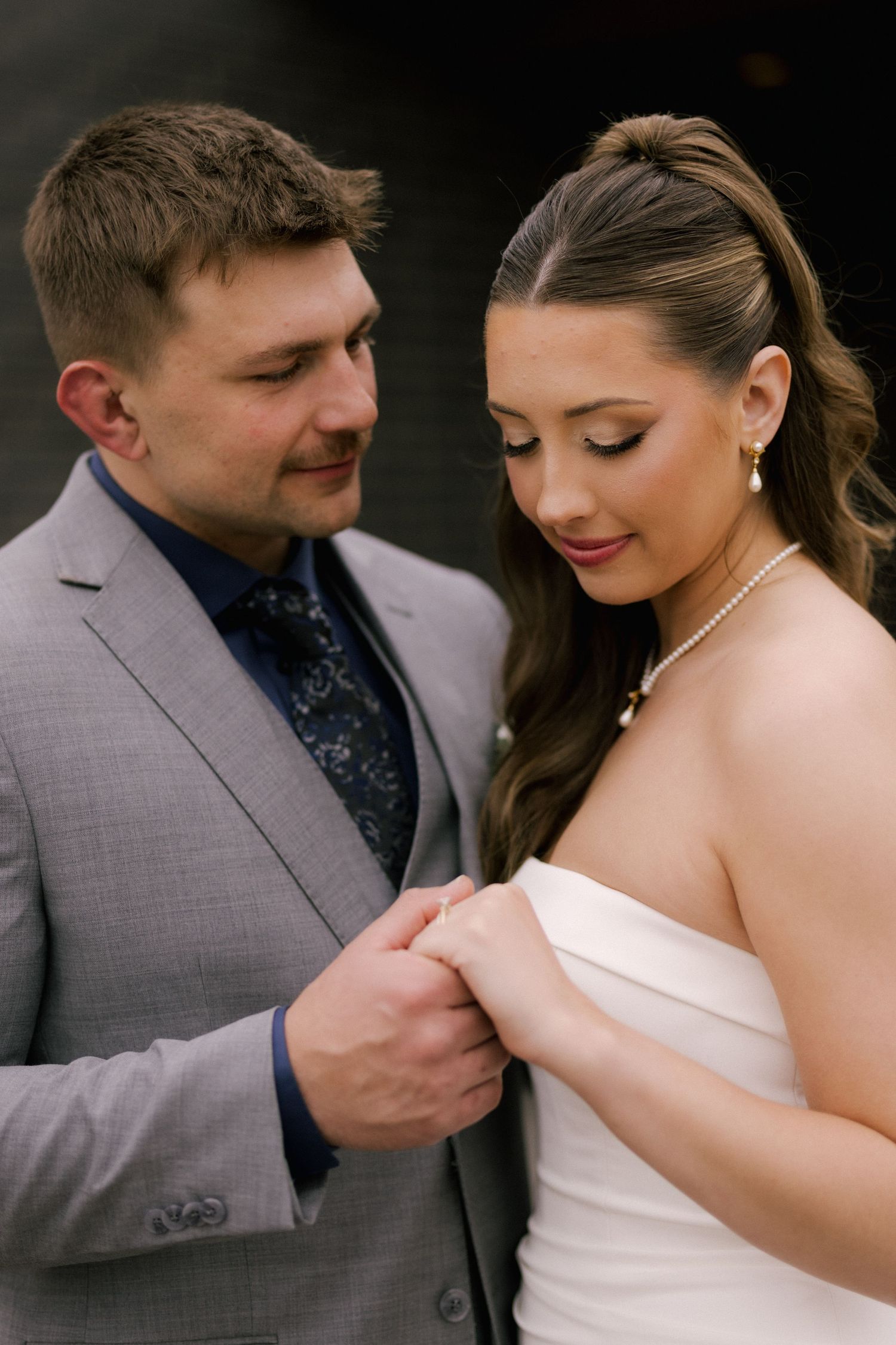 A bride and groom share a tender moment, holding hands, she in a white strapless gown with pearls, he in a gray suit.