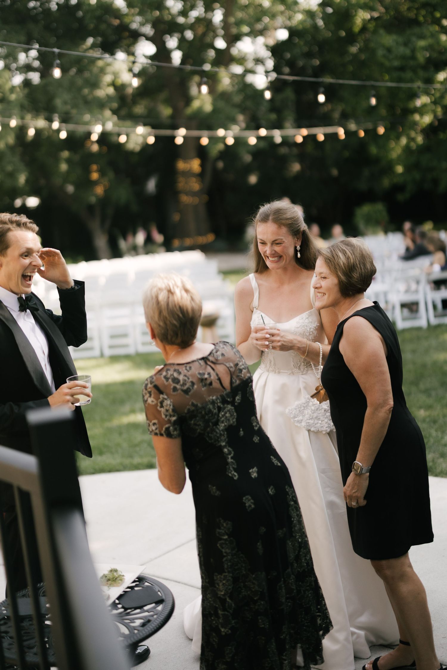 Bride in white gown laughing with guests at outdoor wedding reception with string lights overhead.