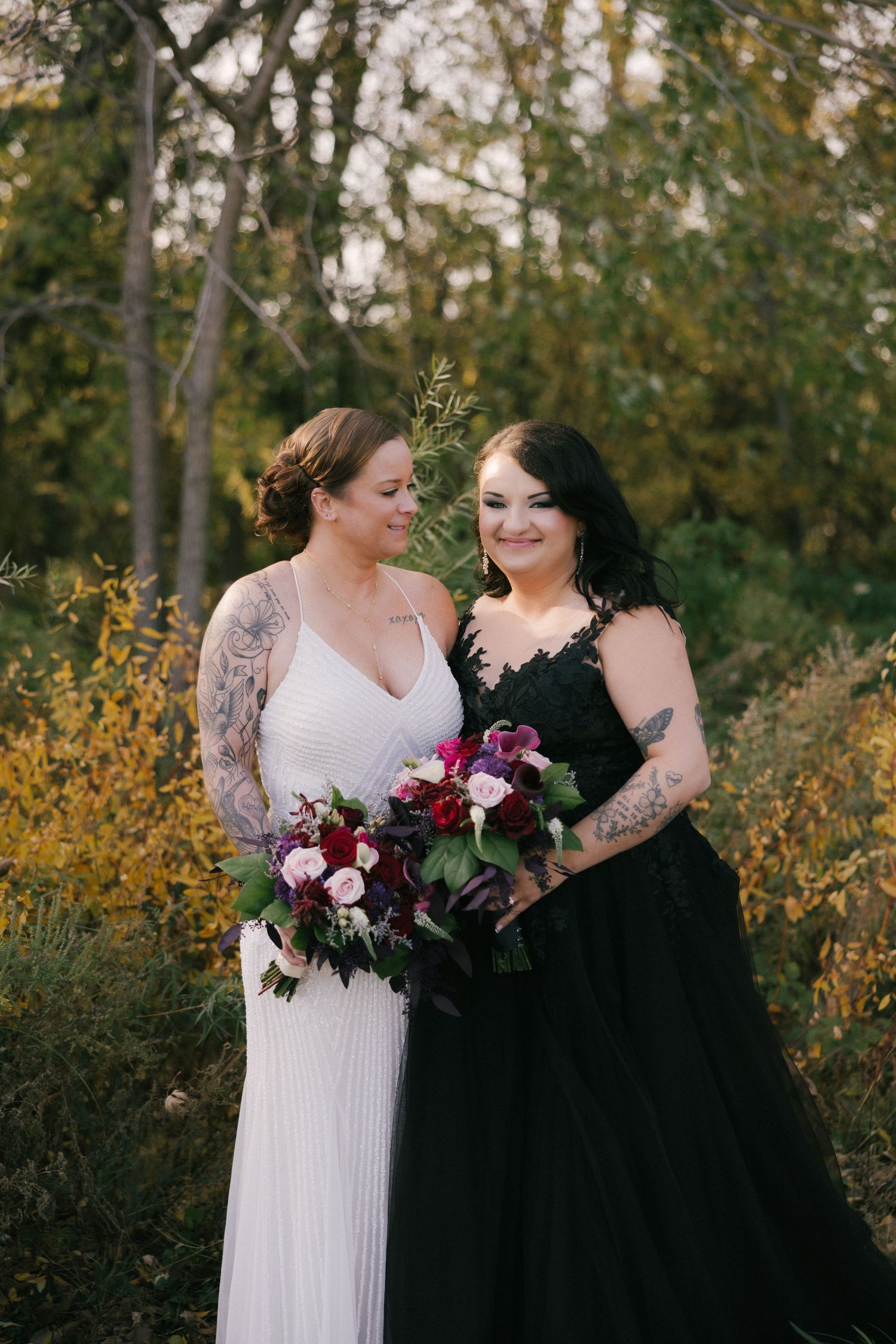 Two people in contrasting white and black formal dresses share a joyful moment surrounded by autumn foliage.