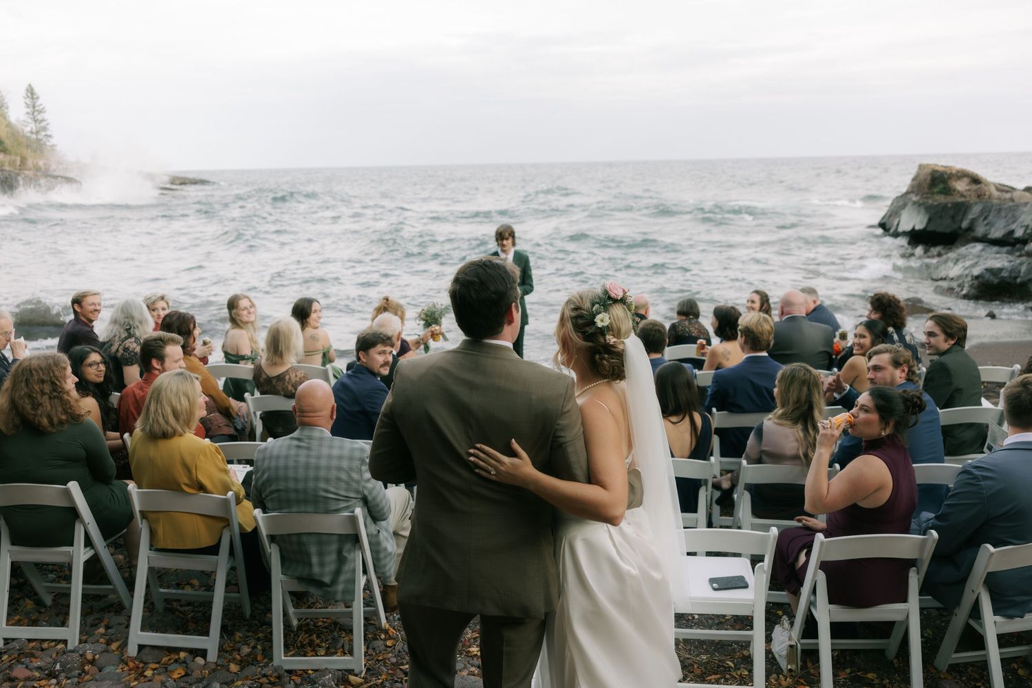 A scenic outdoor wedding ceremony overlooking the ocean with guests seated in white chairs on the rocky coastline.