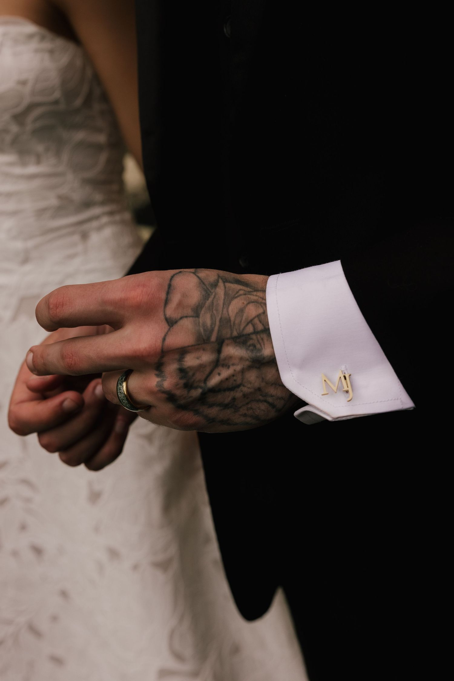 Groom with tattooed hand holding bride's hand, wearing black suit with white cuff and gold MJ monogram cufflink.